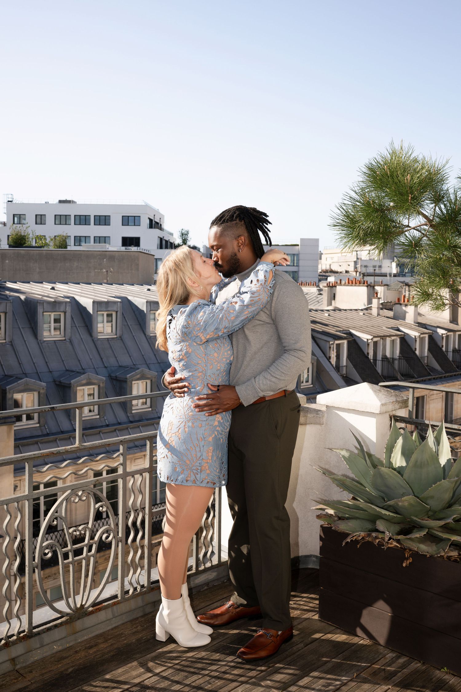 Couple embracing and kissing on Parisian rooftop terrace with classic Haussmann buildings in background.