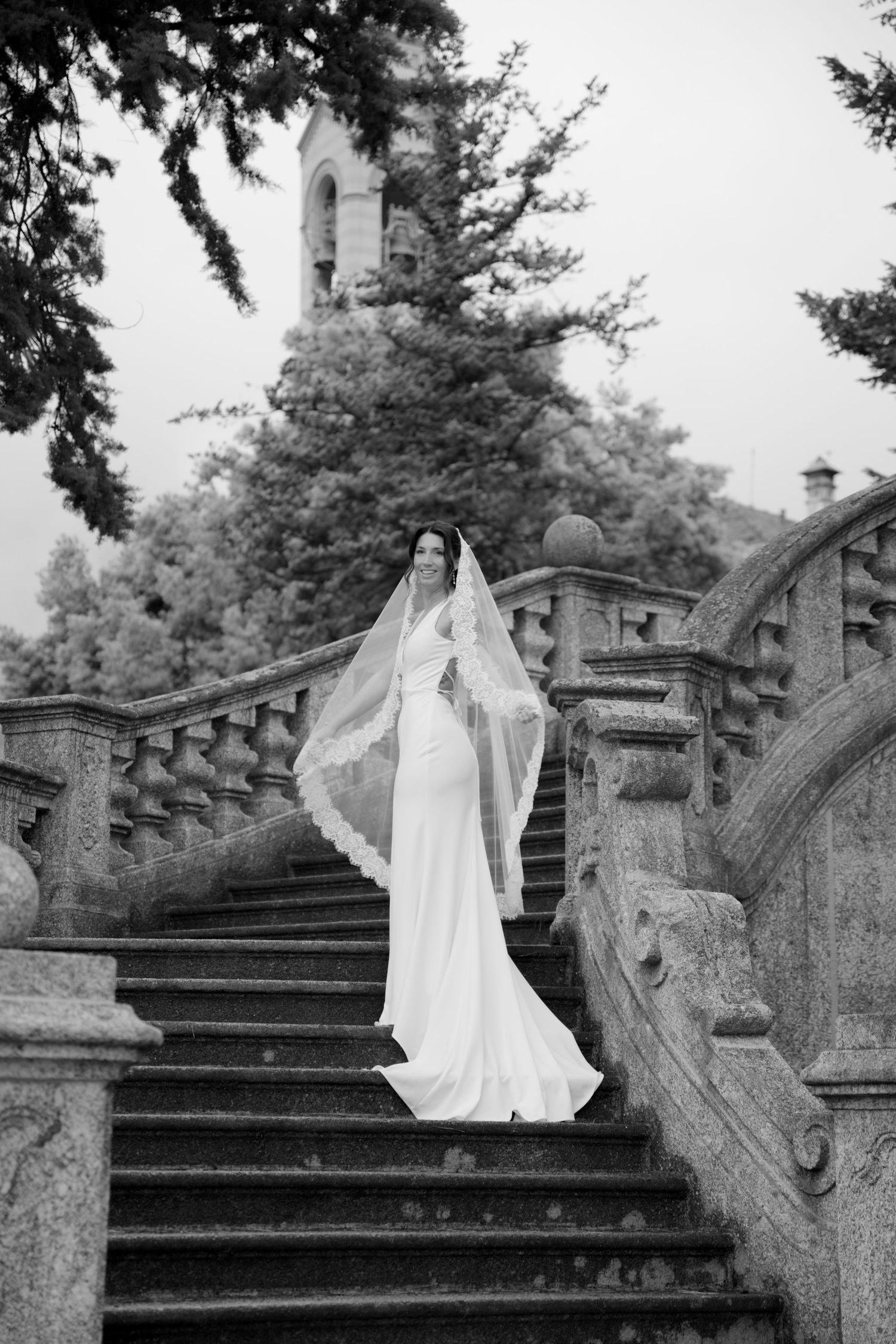 Elegant bride in white gown and veil descending grand stone staircase at historic venue.