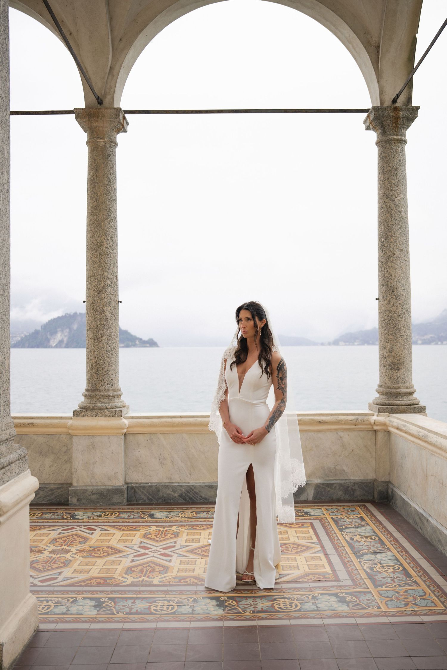 Bride in white slit gown stands under marble arches of Italian villa loggia overlooking Lake Como.