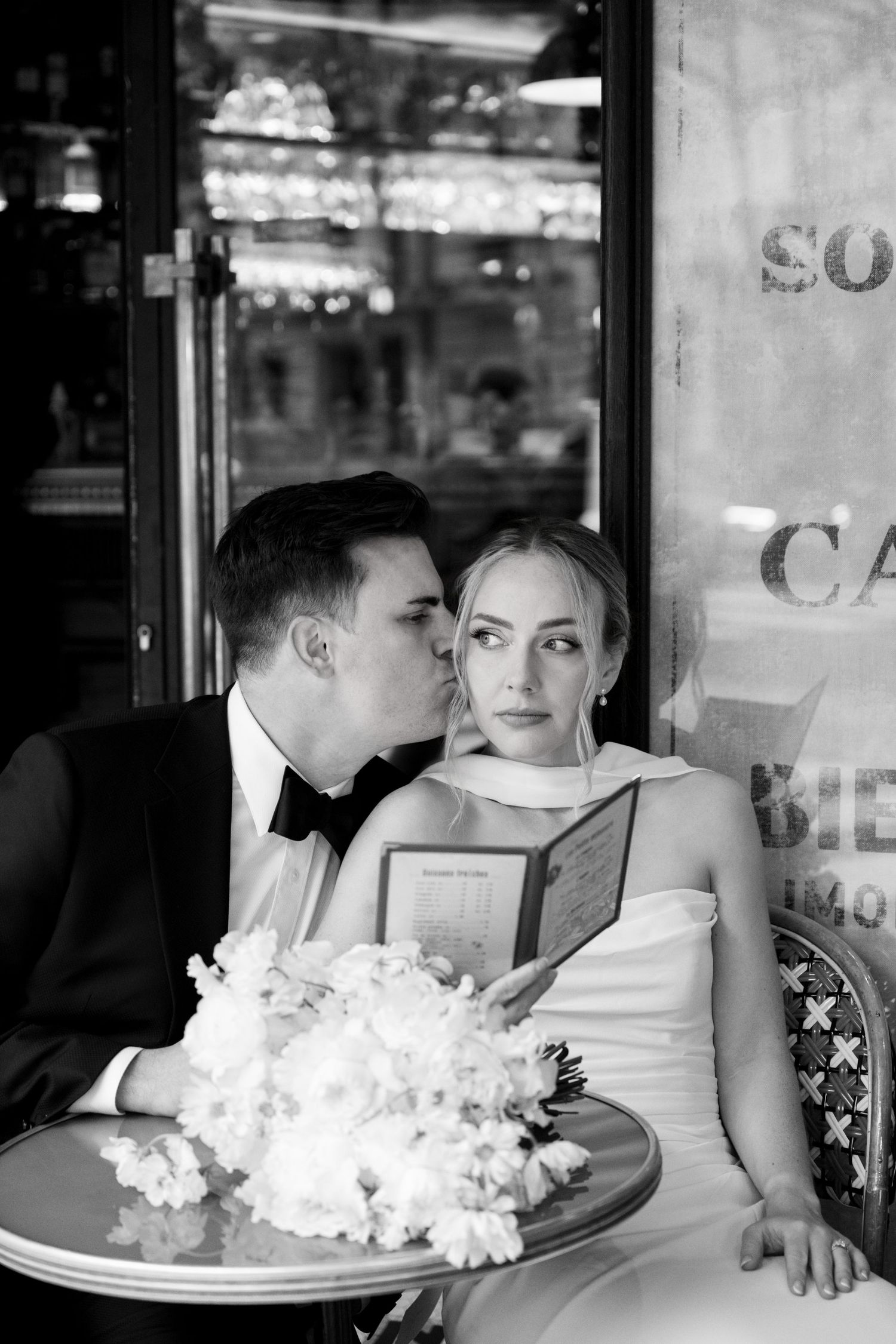 A romantic black and white series of newlyweds sharing intimate moments at a restaurant table with white flowers.