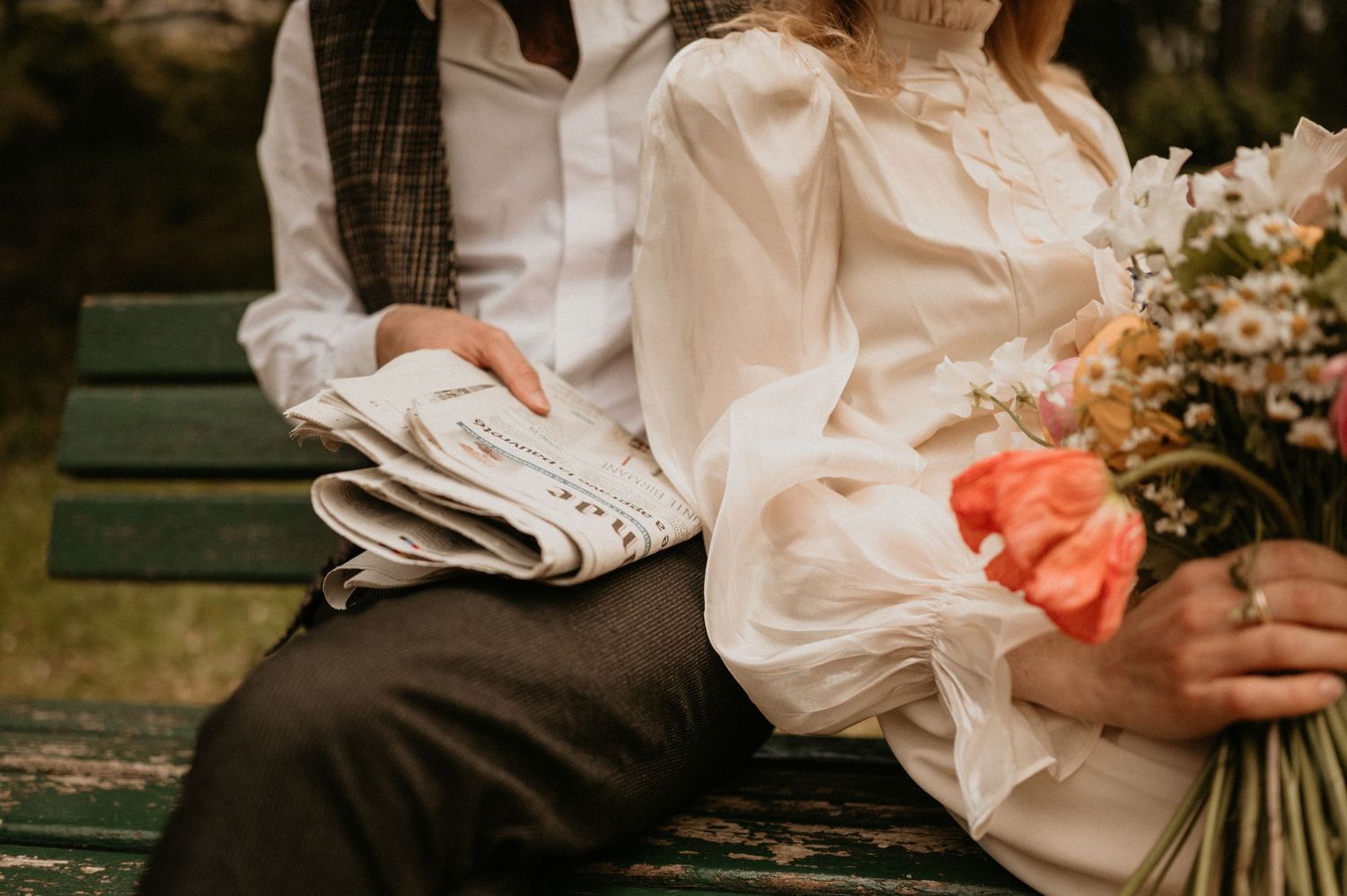 A close-up shot of hands holding a newspaper and flowers while sitting on a park bench.
