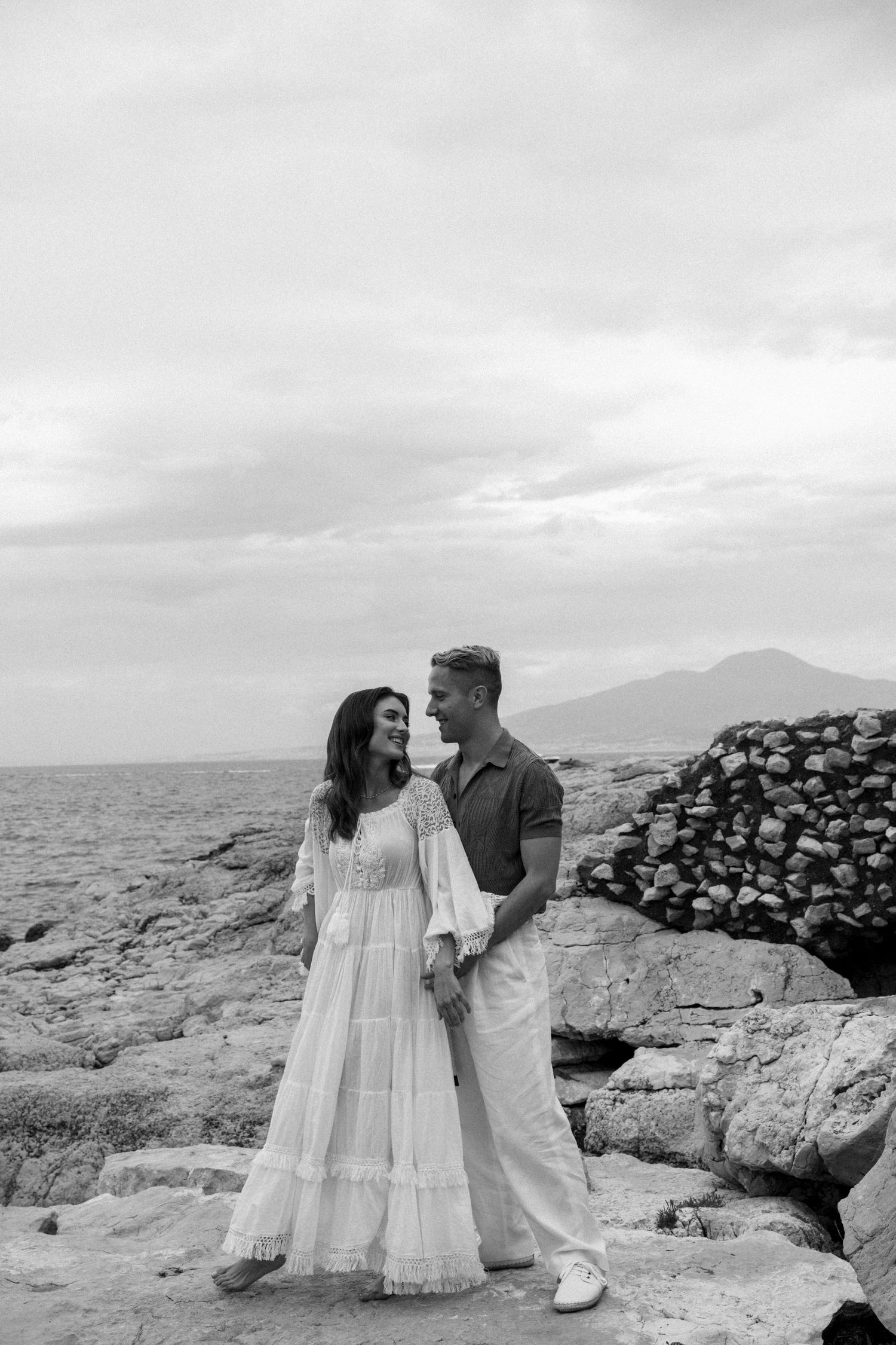 A couple in casual attire poses on rocky coastal terrain with mountains and ocean in the background.