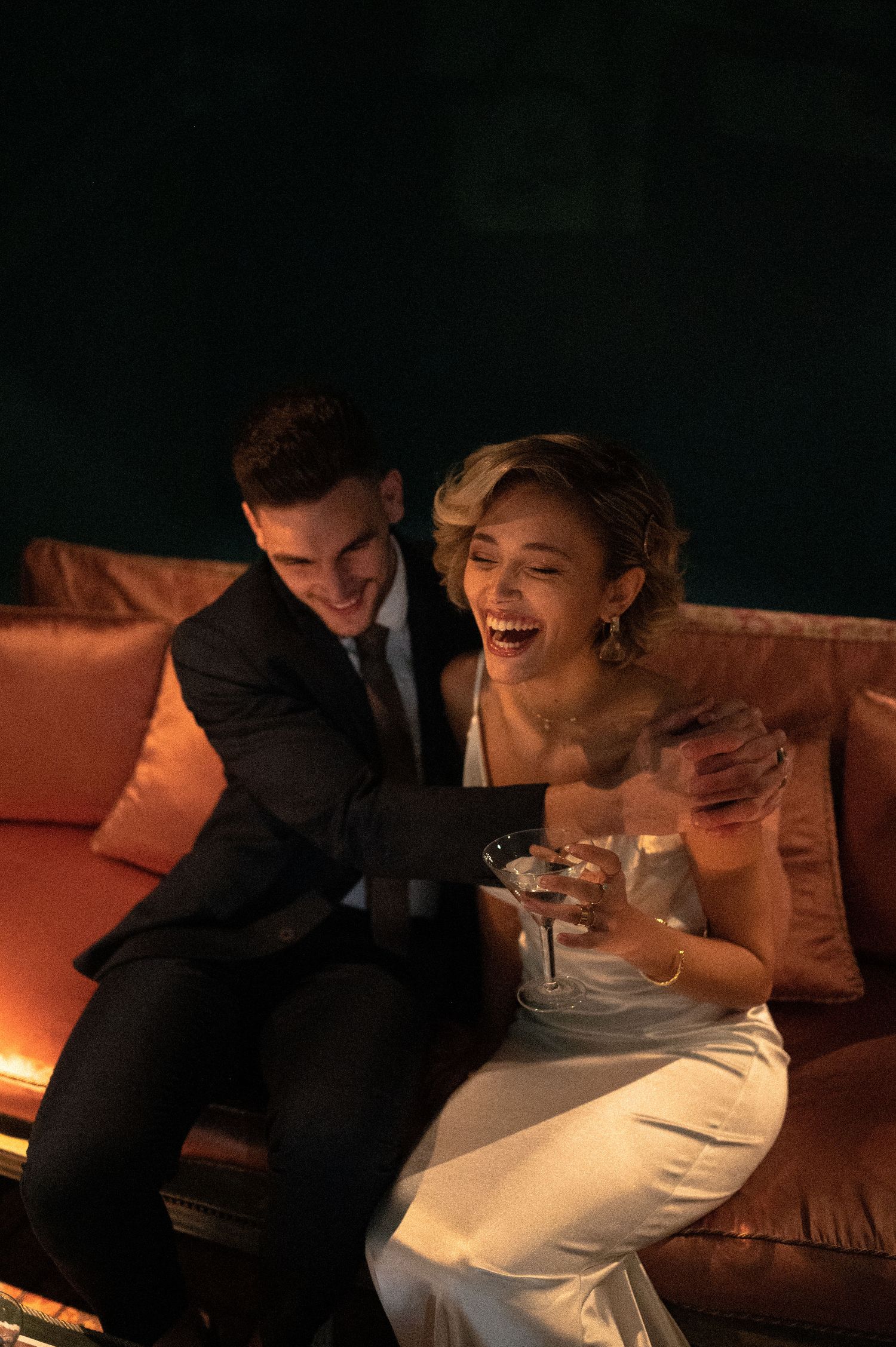 Couple shares joyful moment on orange couch during evening wedding reception.