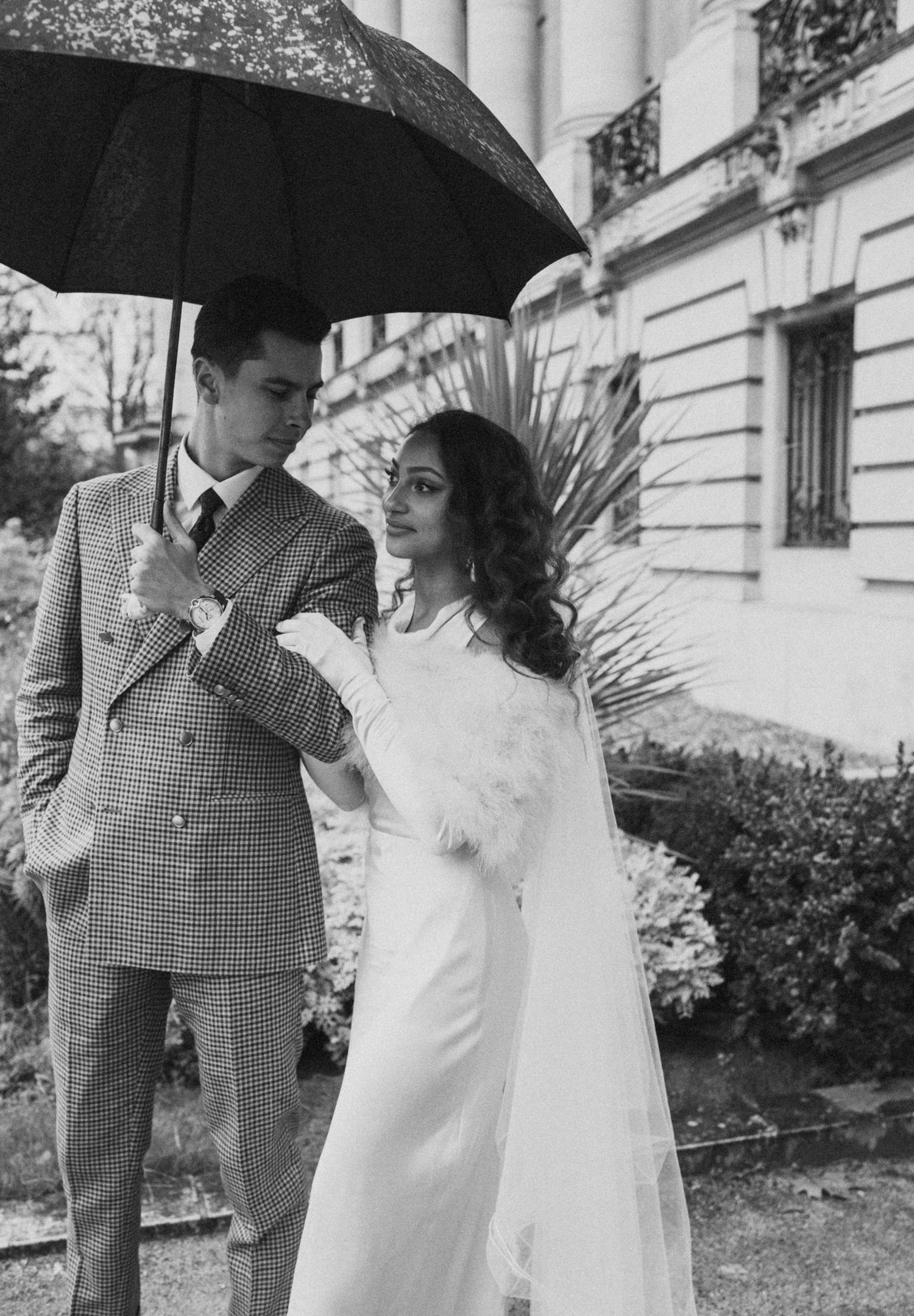 Monochrome portrait of a couple in vintage wedding attire sharing an umbrella in a garden setting.