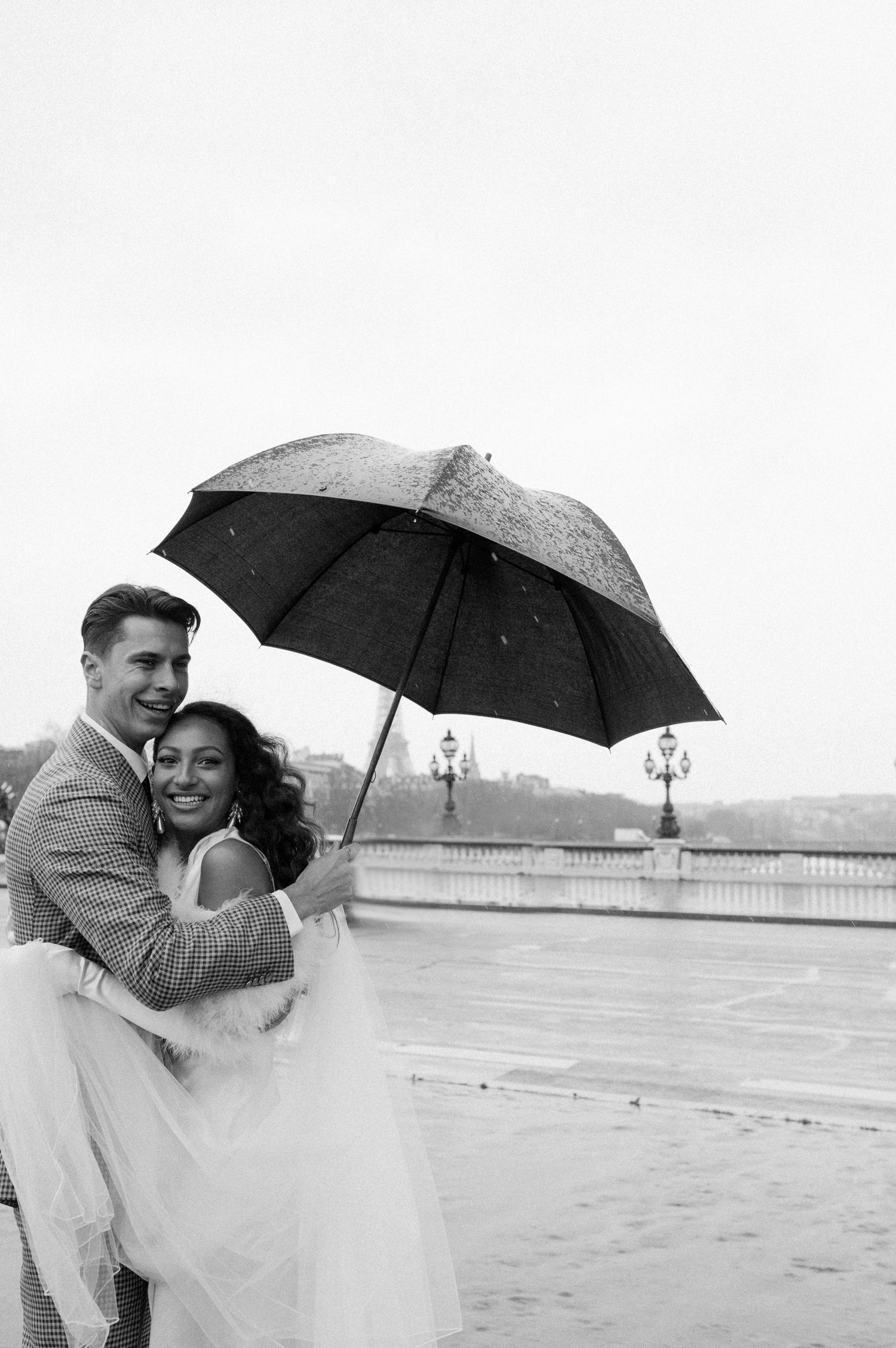 Newlyweds embrace in the rain with Parisian architecture visible in background.