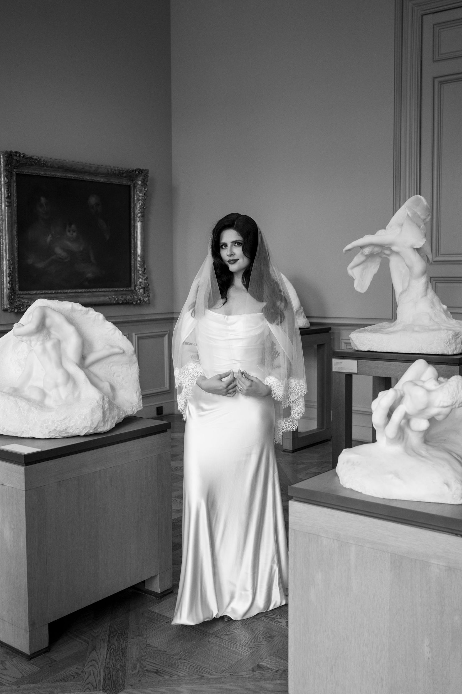Black and white portrait of bride in satin gown and lace veil standing among marble sculptures in gallery.