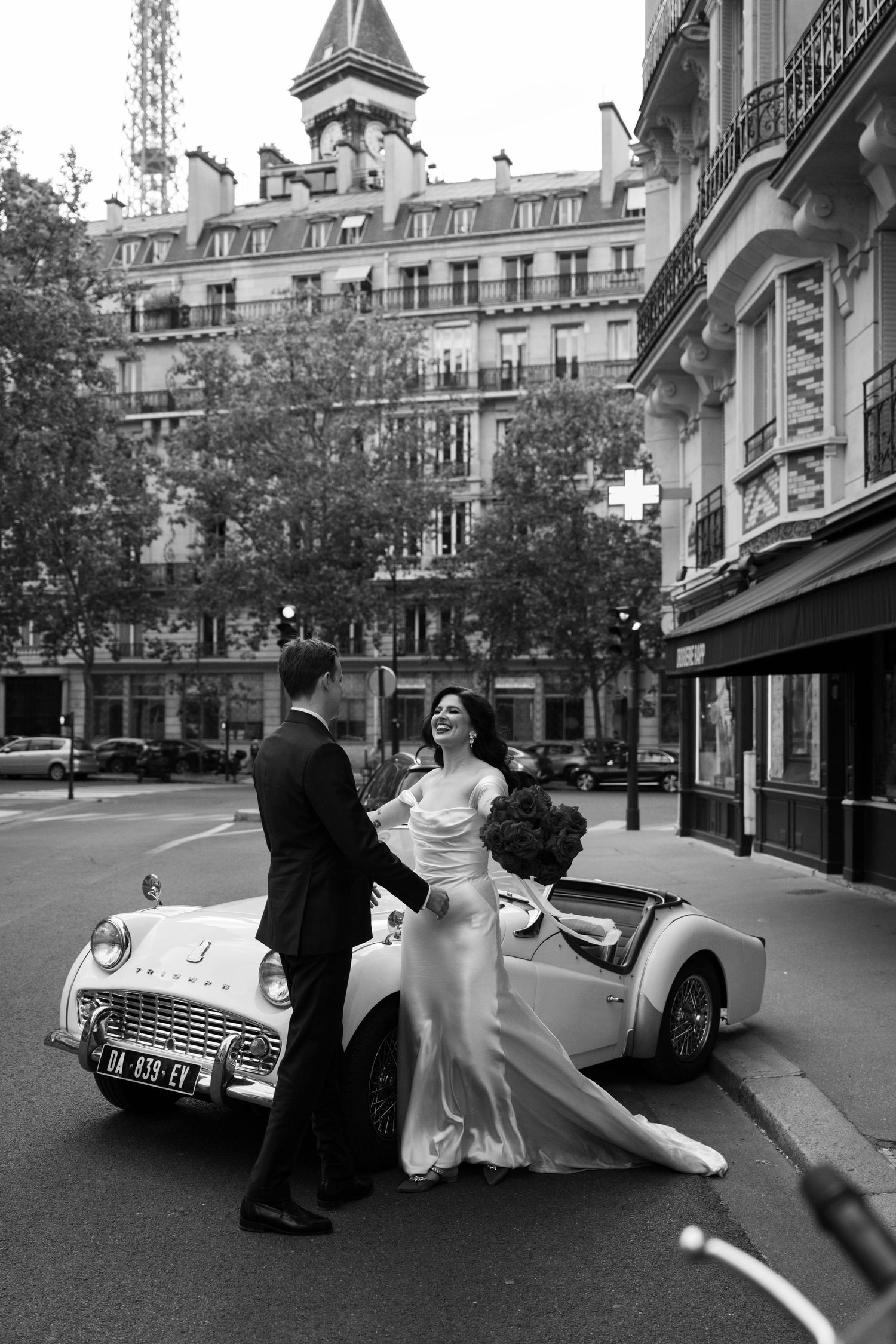 Black and white photo of bride and groom posing beside vintage white convertible with Eiffel Tower backdrop.
