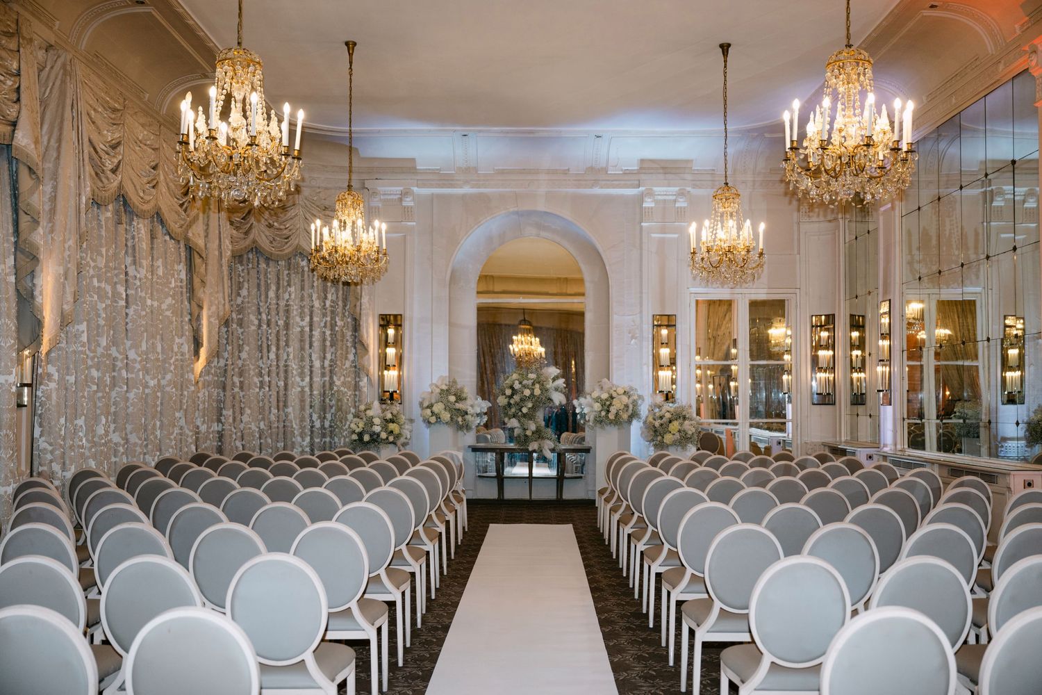 Luxurious wedding ceremony room featuring ornate gold chandeliers, grey upholstered chairs, and a white floral altar.