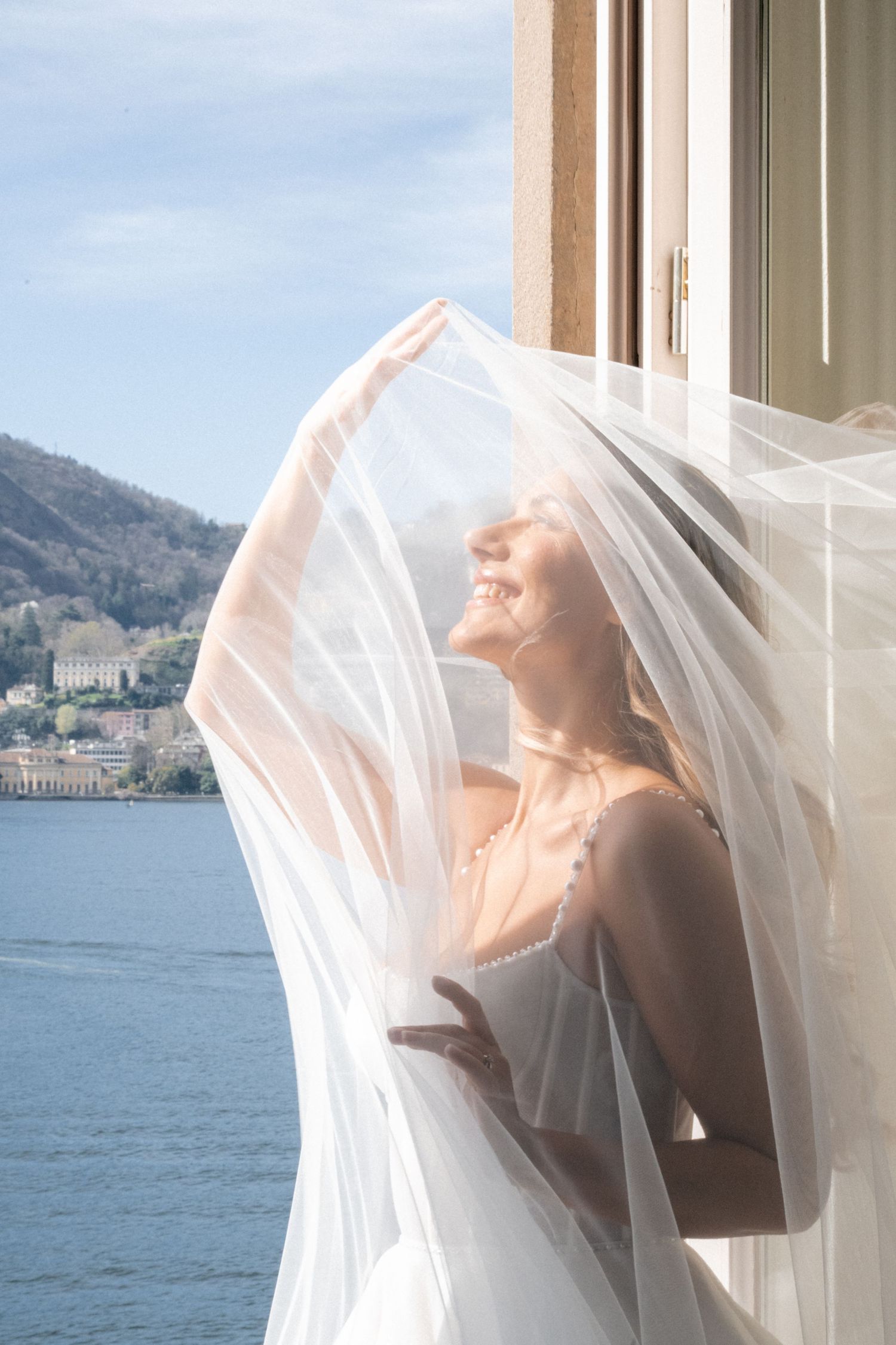 Radiant bride lifting veil in sunlight at open window overlooking Lake Como waterfront.