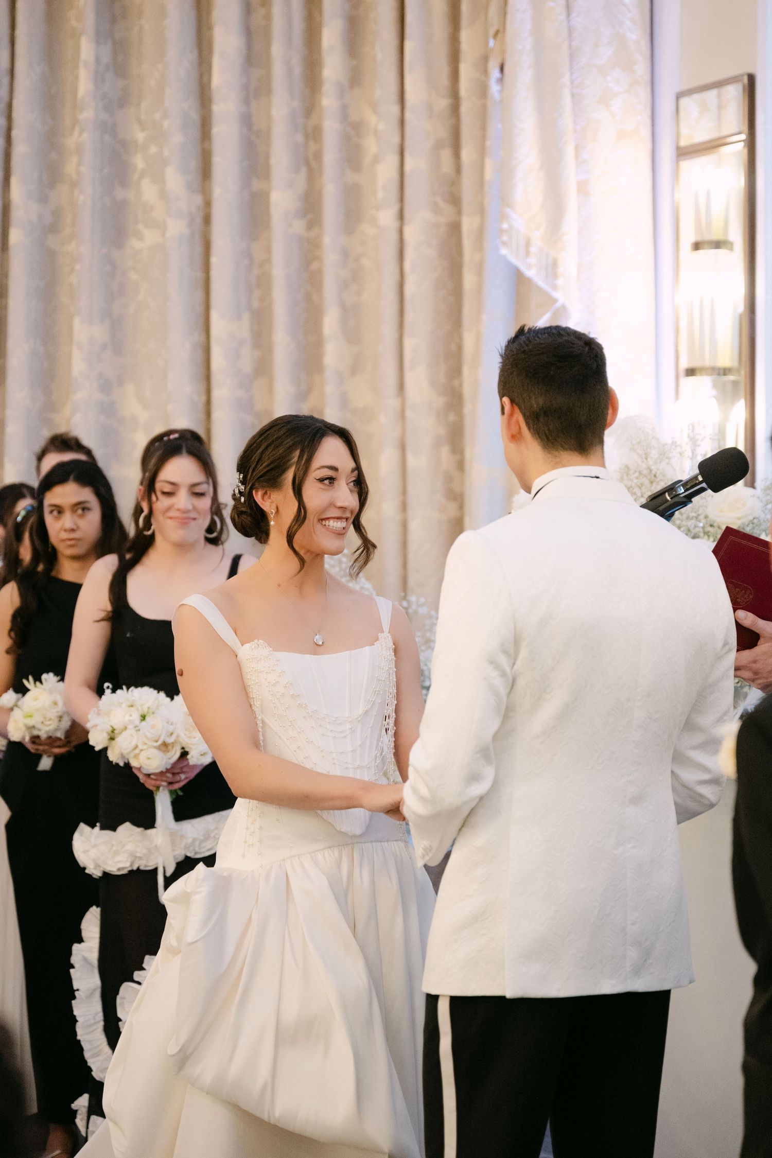 Bride smiling at groom during indoor wedding ceremony with bridesmaids in black dresses holding white bouquets.