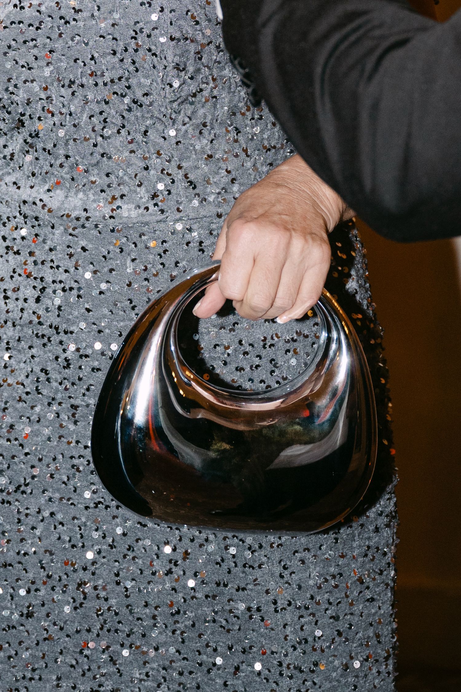 Close-up of guest holding sleek metallic dark chrome handbag against sparkling silver sequined dress at formal event.
