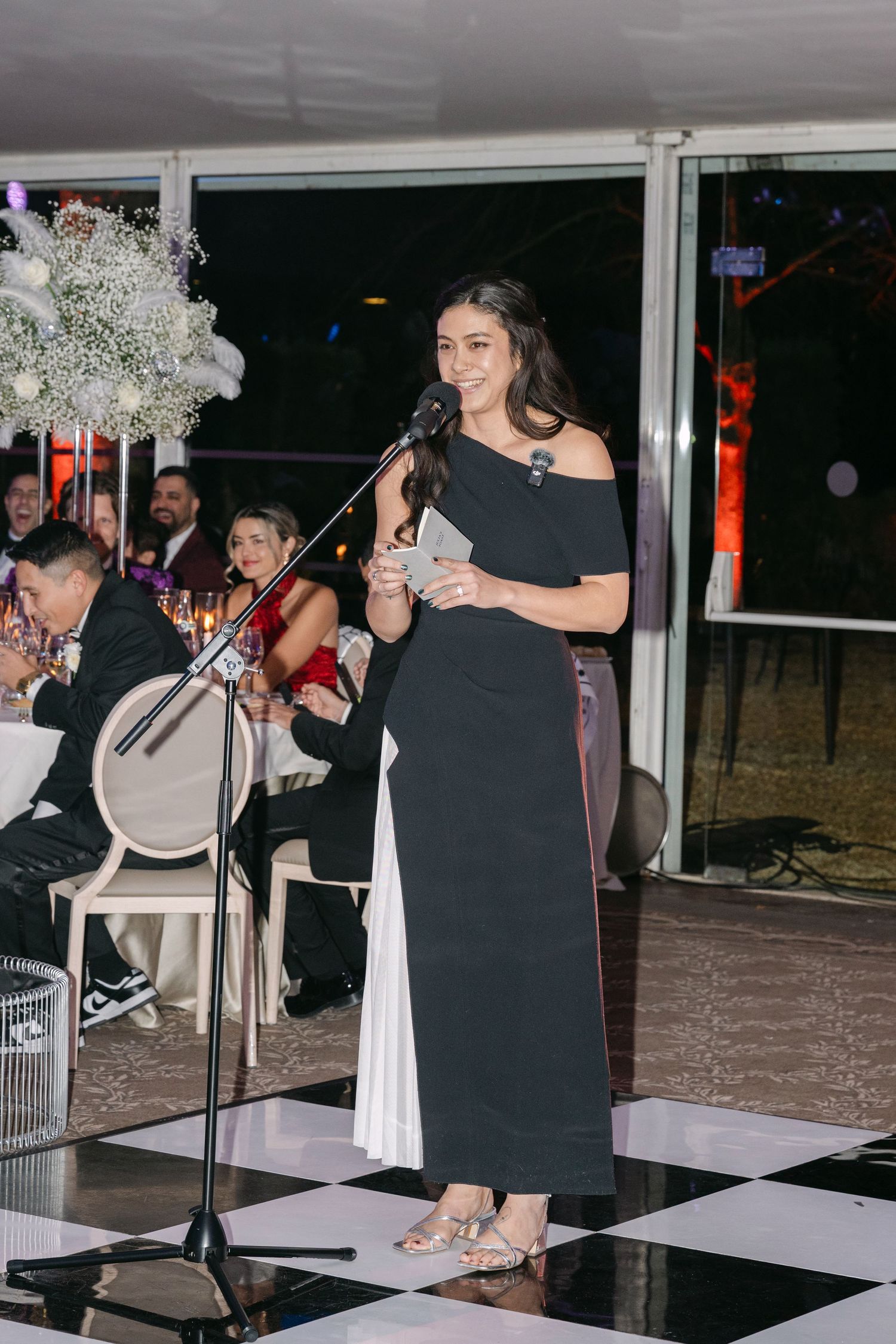 Elegant woman in black gown delivers speech at microphone during wedding reception ceremony.