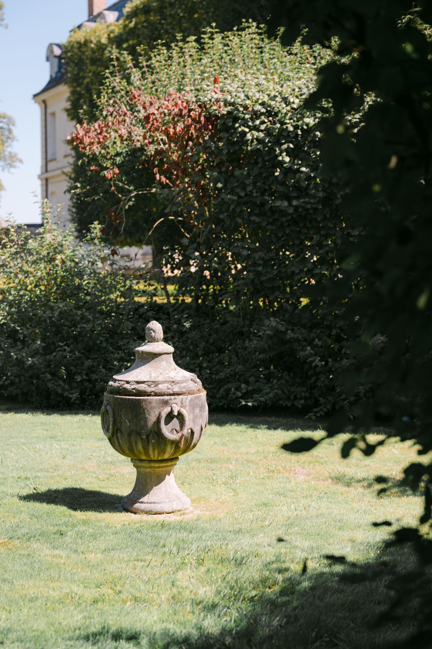 Ornate stone garden urn on green lawn surrounded by lush hedges and flowering shrubs at a French château.