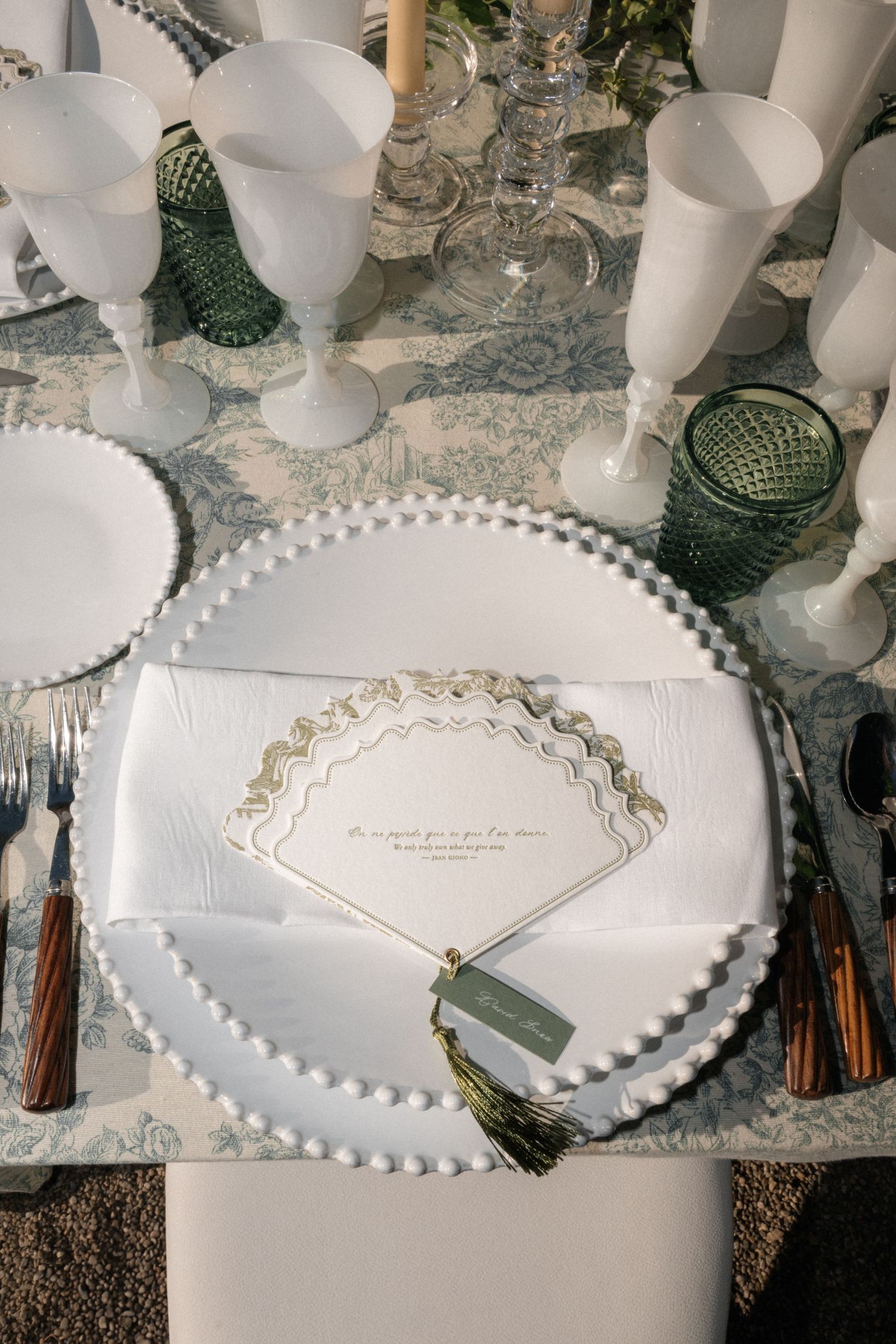 Elegant outdoor place setting with beaded white plate, menu card, tassel, and green glassware.