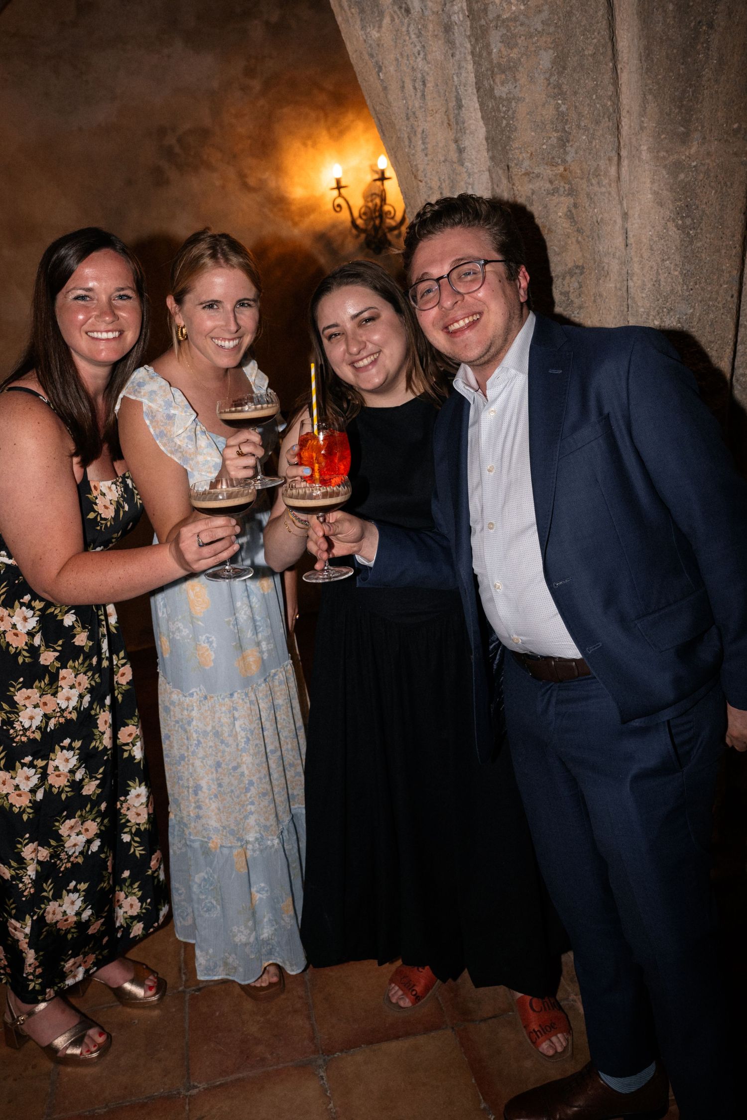Four wedding guests smile and clink cocktail glasses together during an indoor wedding reception celebration.