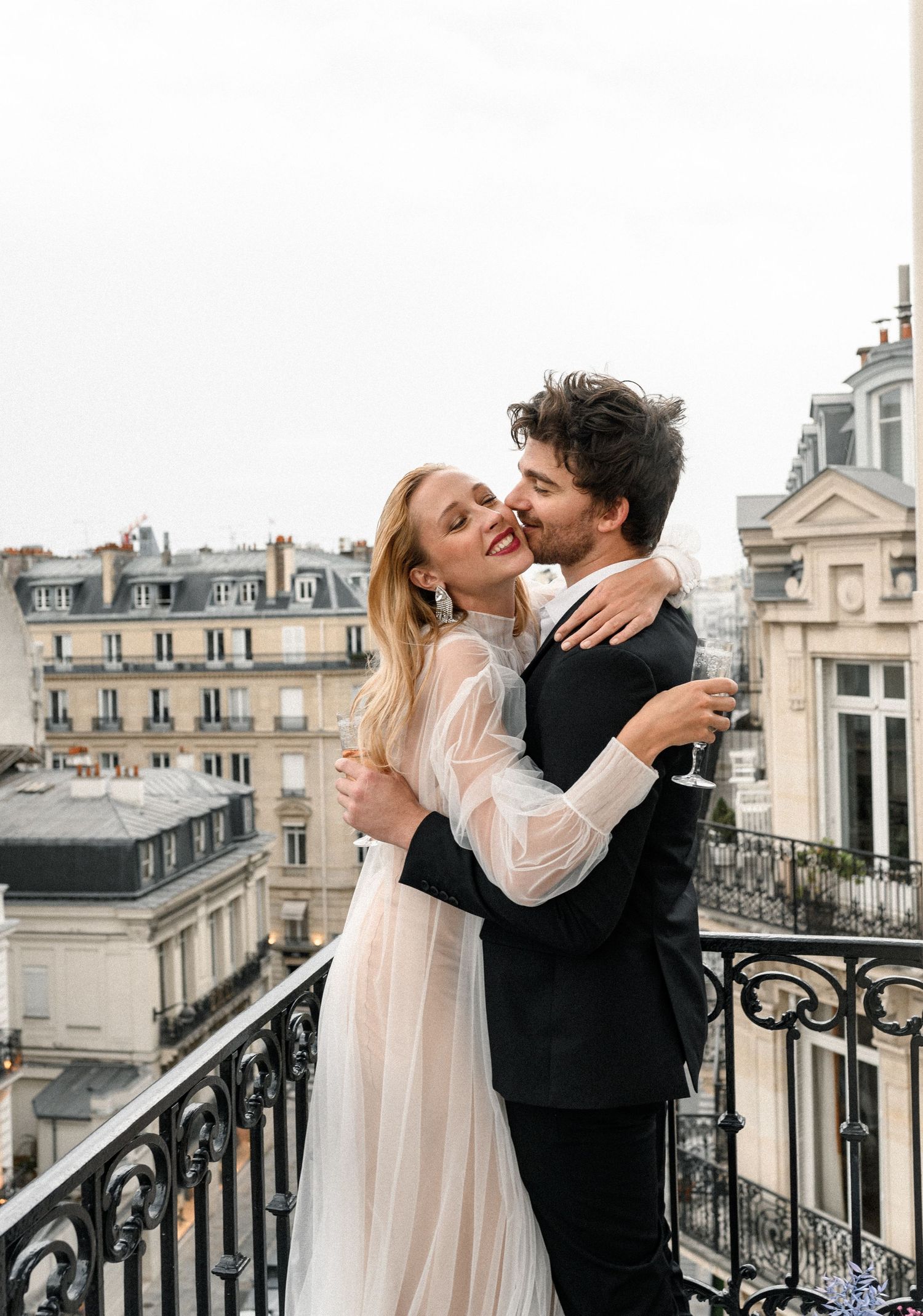 Joyful newlyweds embracing and laughing on a Parisian balcony with Haussmann buildings behind.