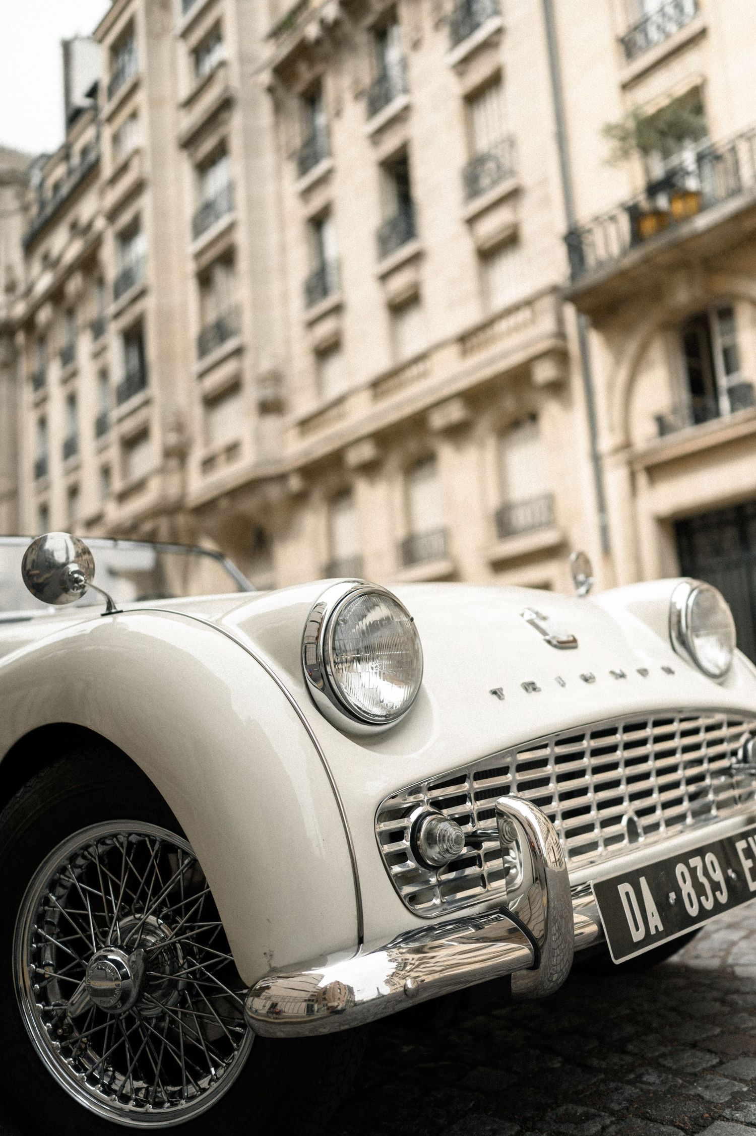 Vintage white Triumph TR3 classic car parked on cobblestone Paris street with elegant Haussmann building backdrop.