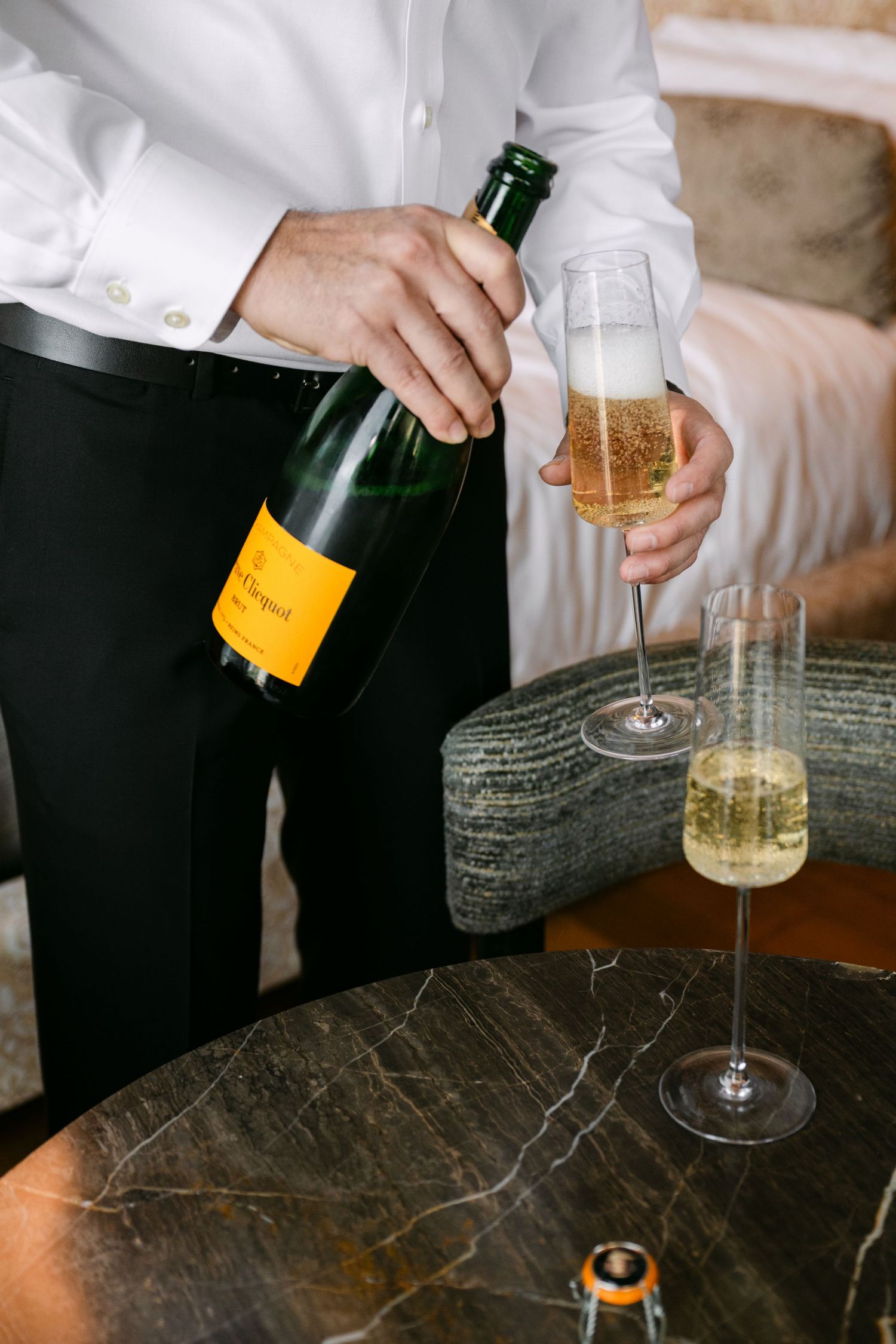 Man in white dress shirt pouring Veuve Clicquot champagne into glasses on a marble table.