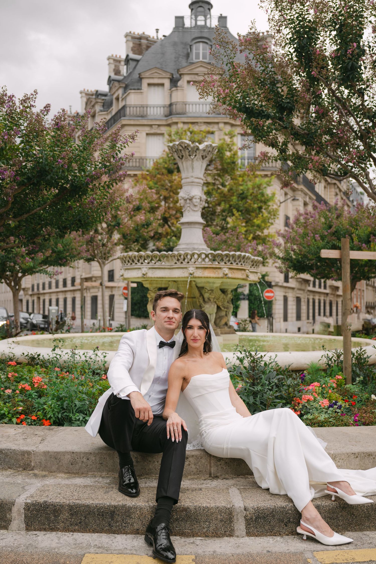 Bride and groom sit together on fountain steps in front of a classic Parisian building with gardens.