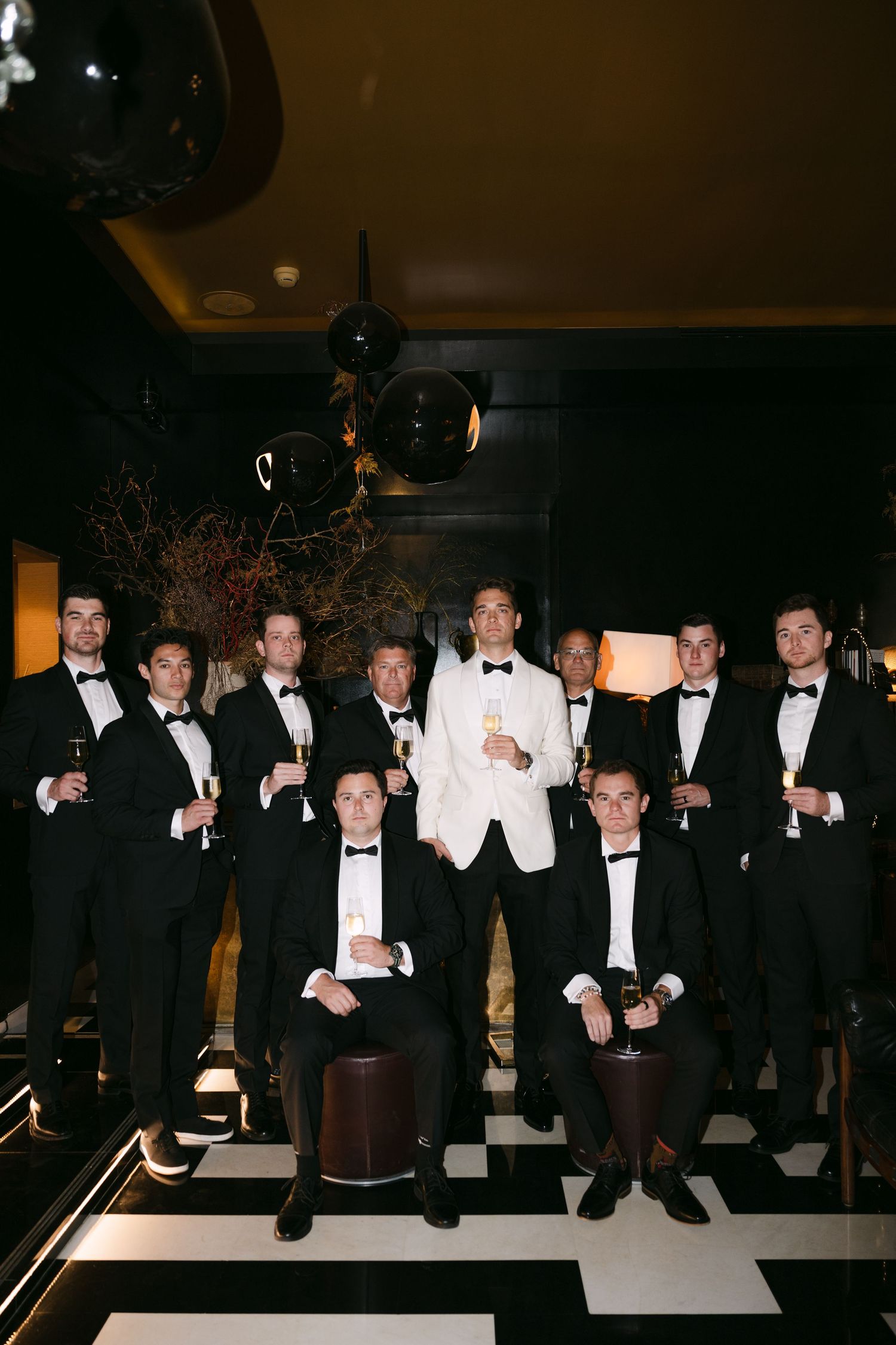 Groomsmen in black tuxedos pose with groom in white jacket on checkered floor at elegant wedding venue.