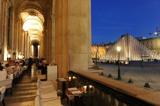 Elegant outdoor dining area under illuminated stone arches with view of the Louvre at night.
