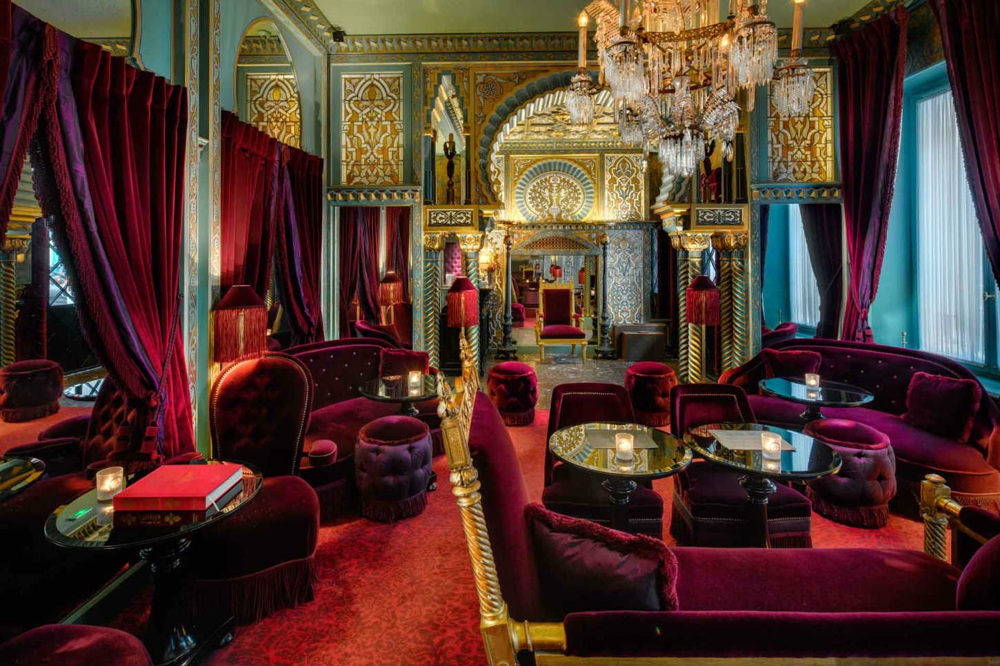 Opulent Moroccan-inspired lounge with rich crimson velvet sofas, gold accents, and ornate chandelier.