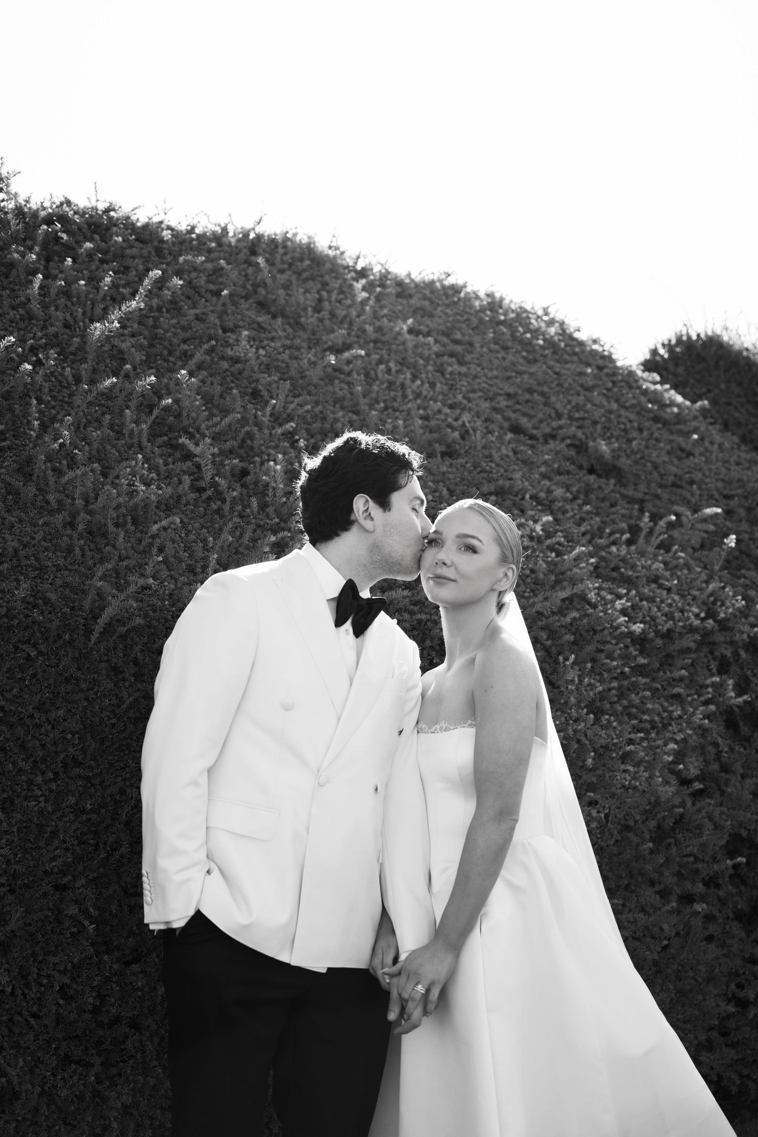 Groom in white tuxedo kisses bride in strapless gown on cheek while holding hands in black and white photo.