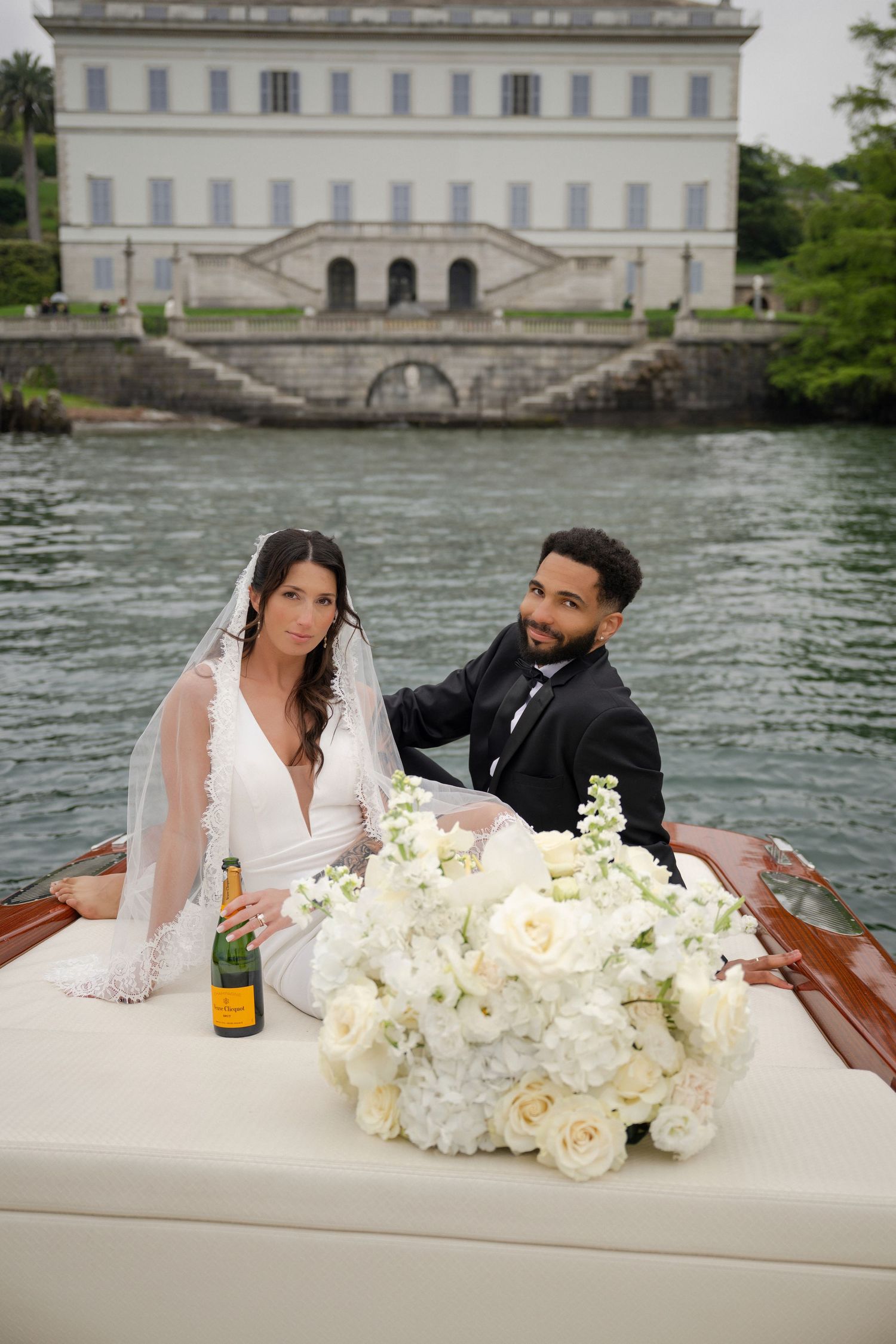 Bride and groom sit on classic wooden boat with white floral bouquet, grand villa reflected in calm lake waters.