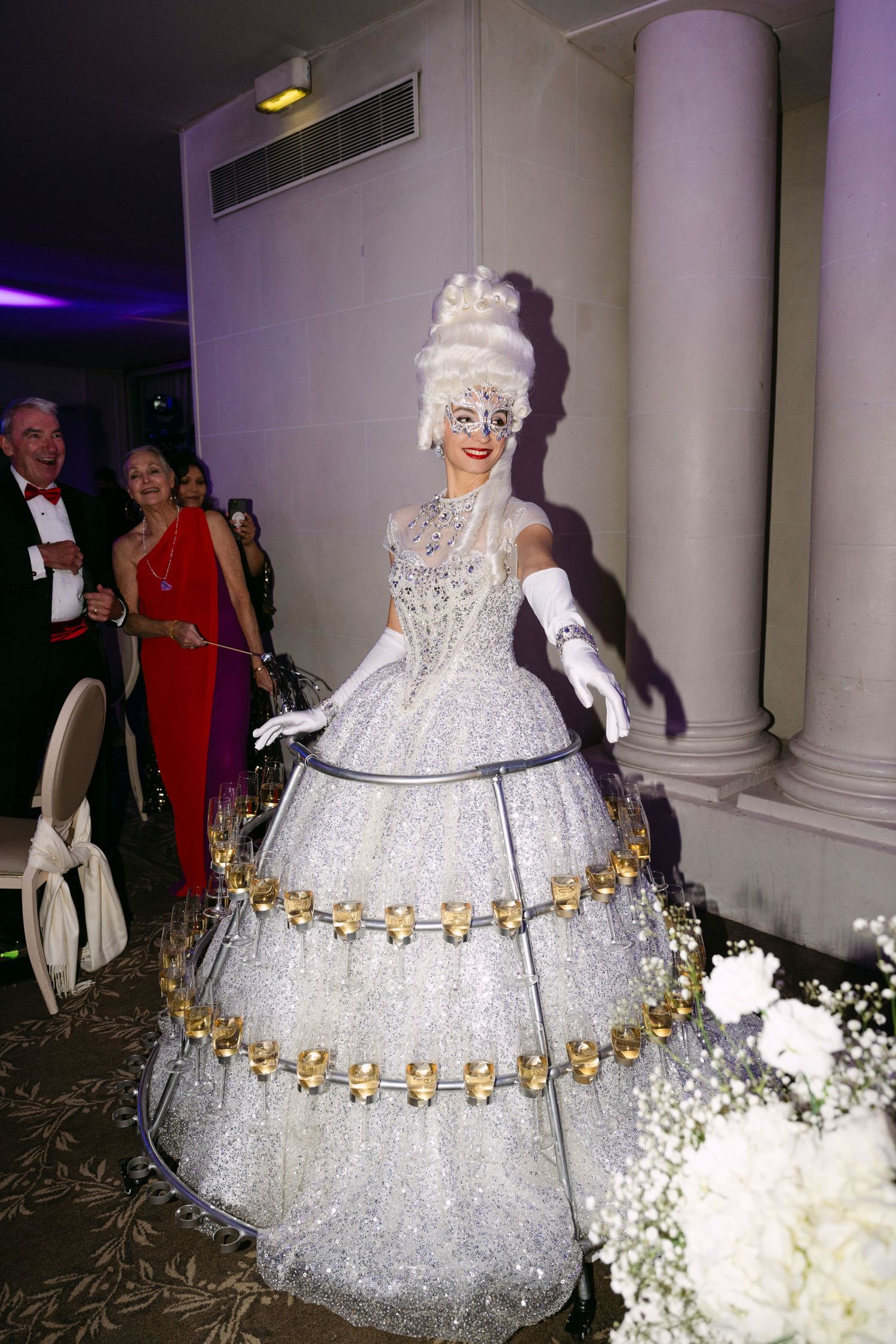 Performer in sparkling white hoop skirt gown with champagne glasses serves drinks at wedding.