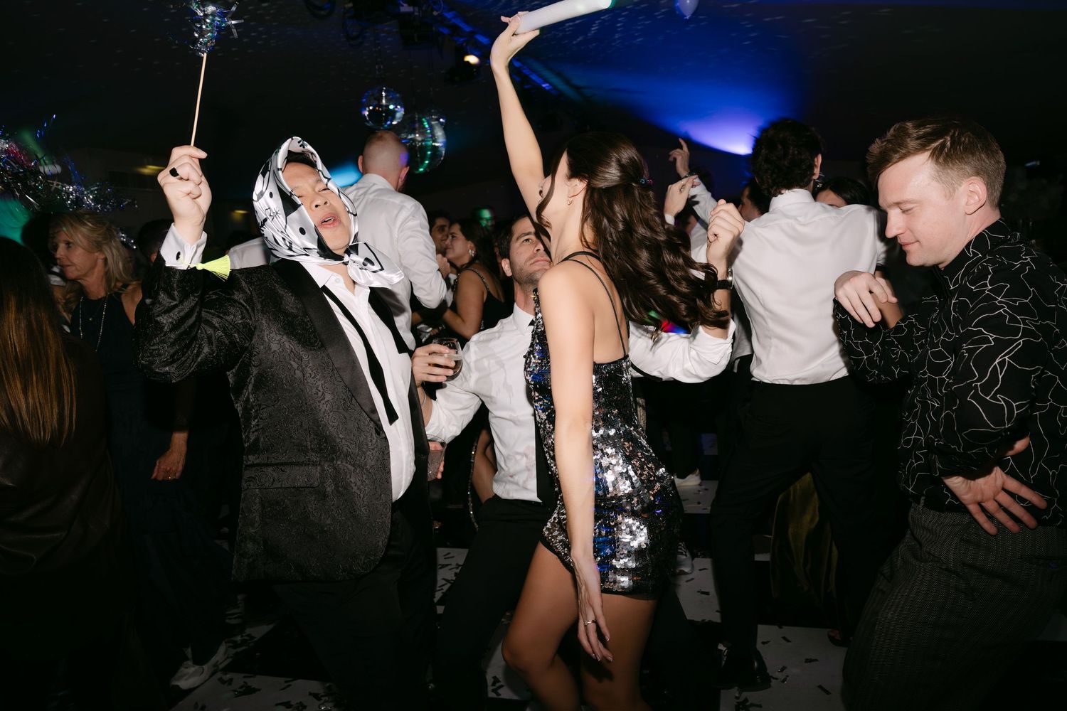 Guests dancing energetically at wedding reception party with confetti and glow sticks on dark dance floor.