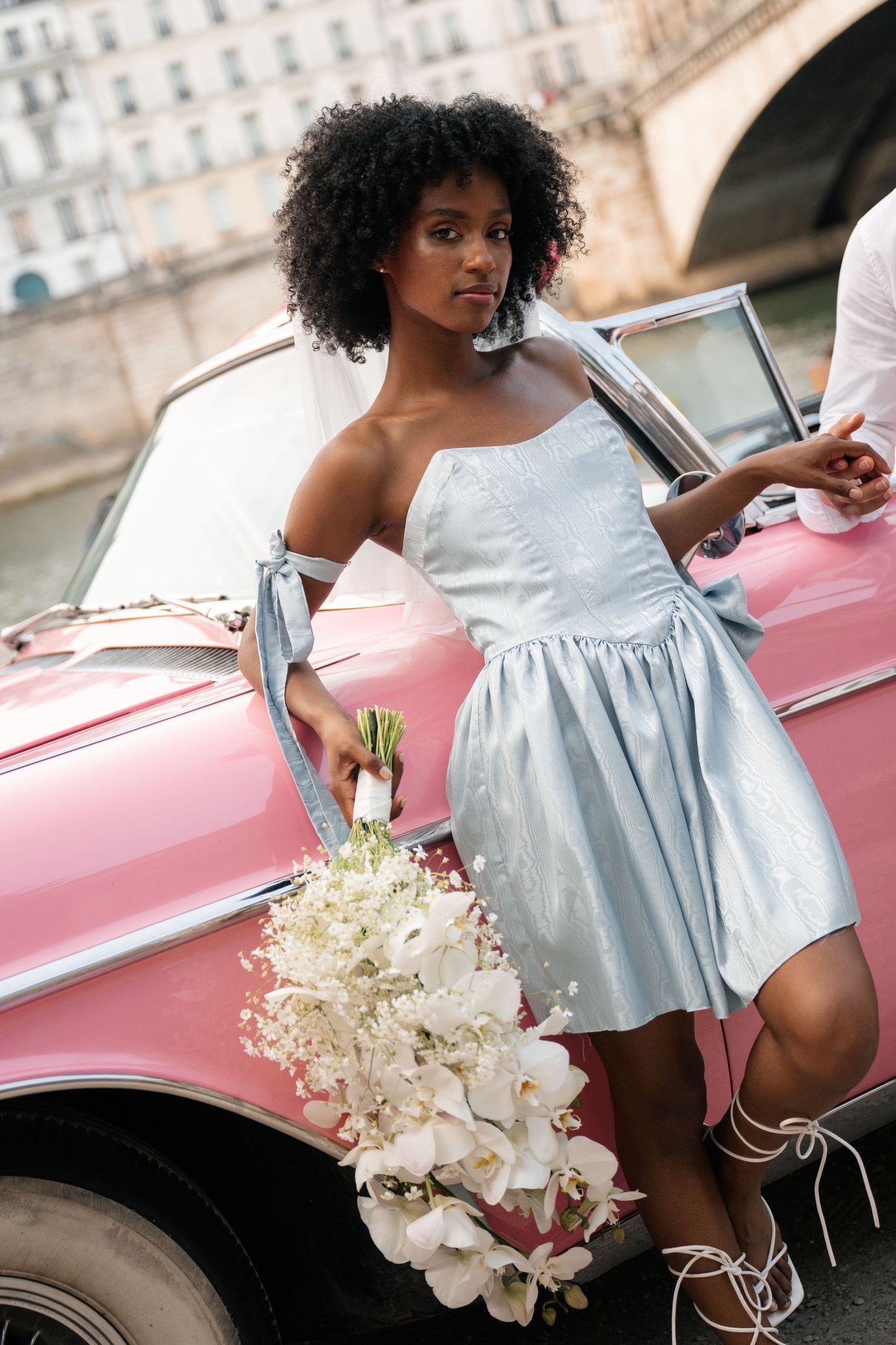 Bride in light blue mini dress holds white orchid bouquet beside vintage pink convertible in Paris.