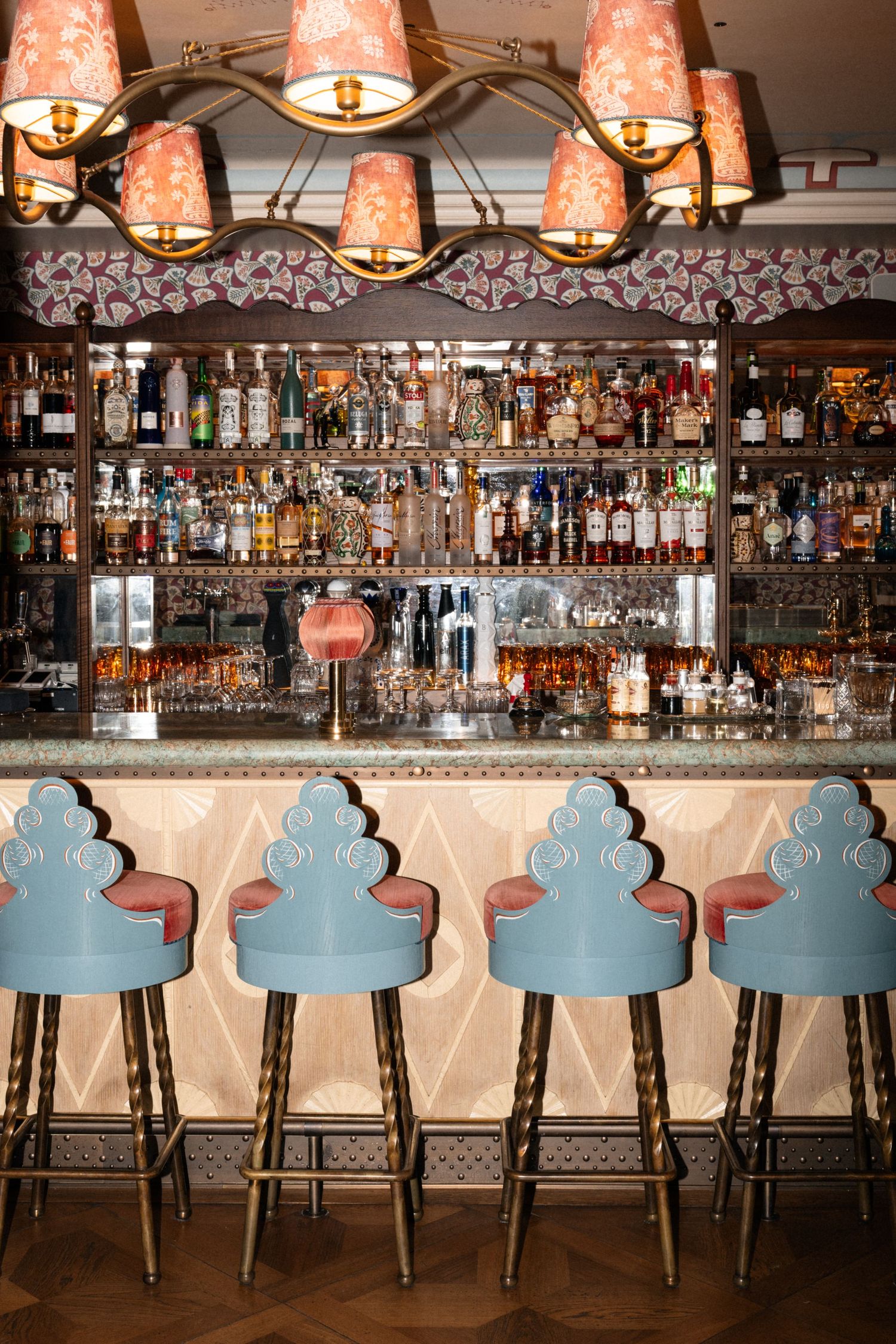 Stylish hotel bar with blue scalloped stools, pink pendant lights, and full liquor shelves.