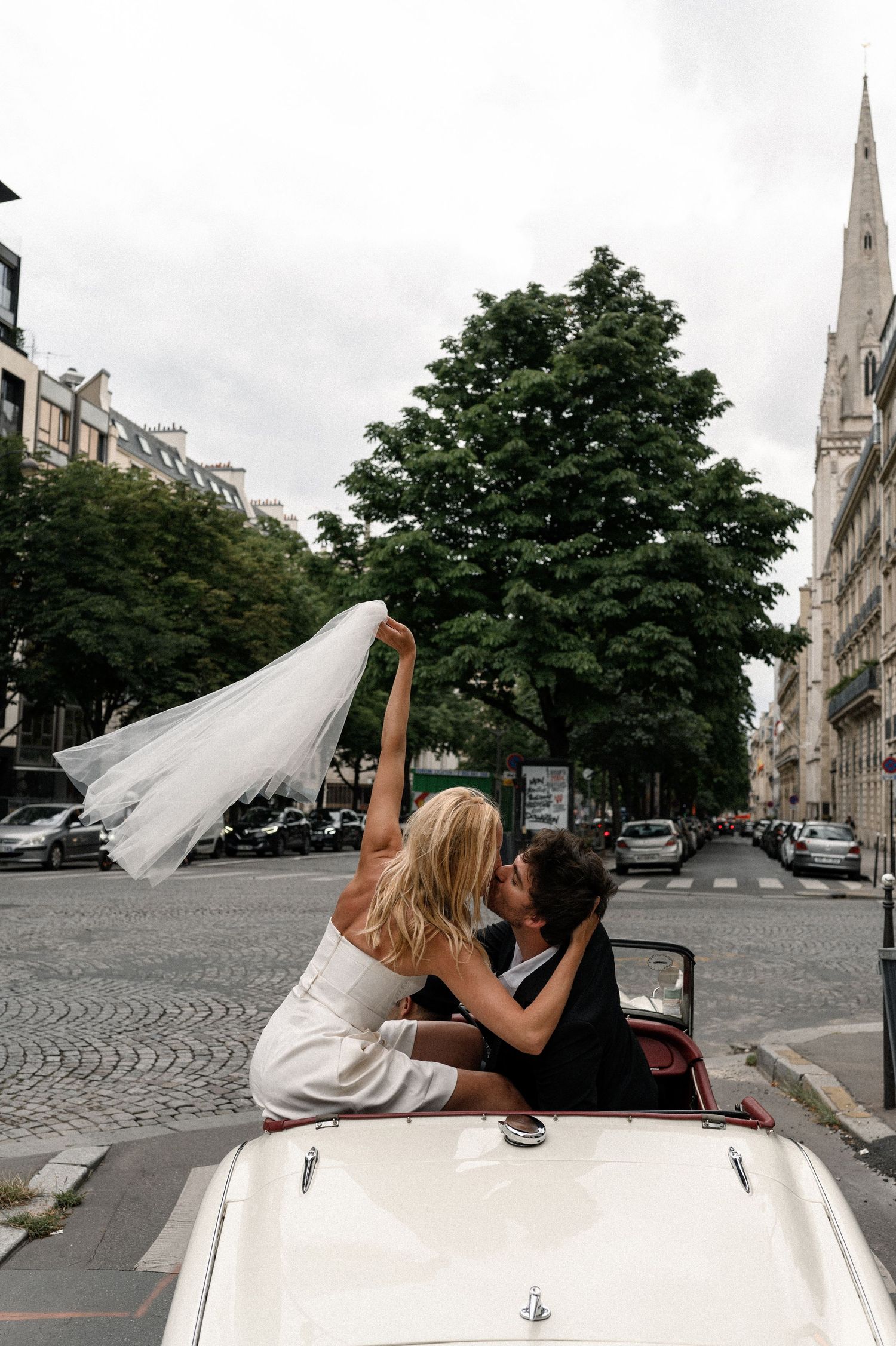 Bride and groom kissing in a vintage white convertible on a Parisian street, veil blowing in the wind.