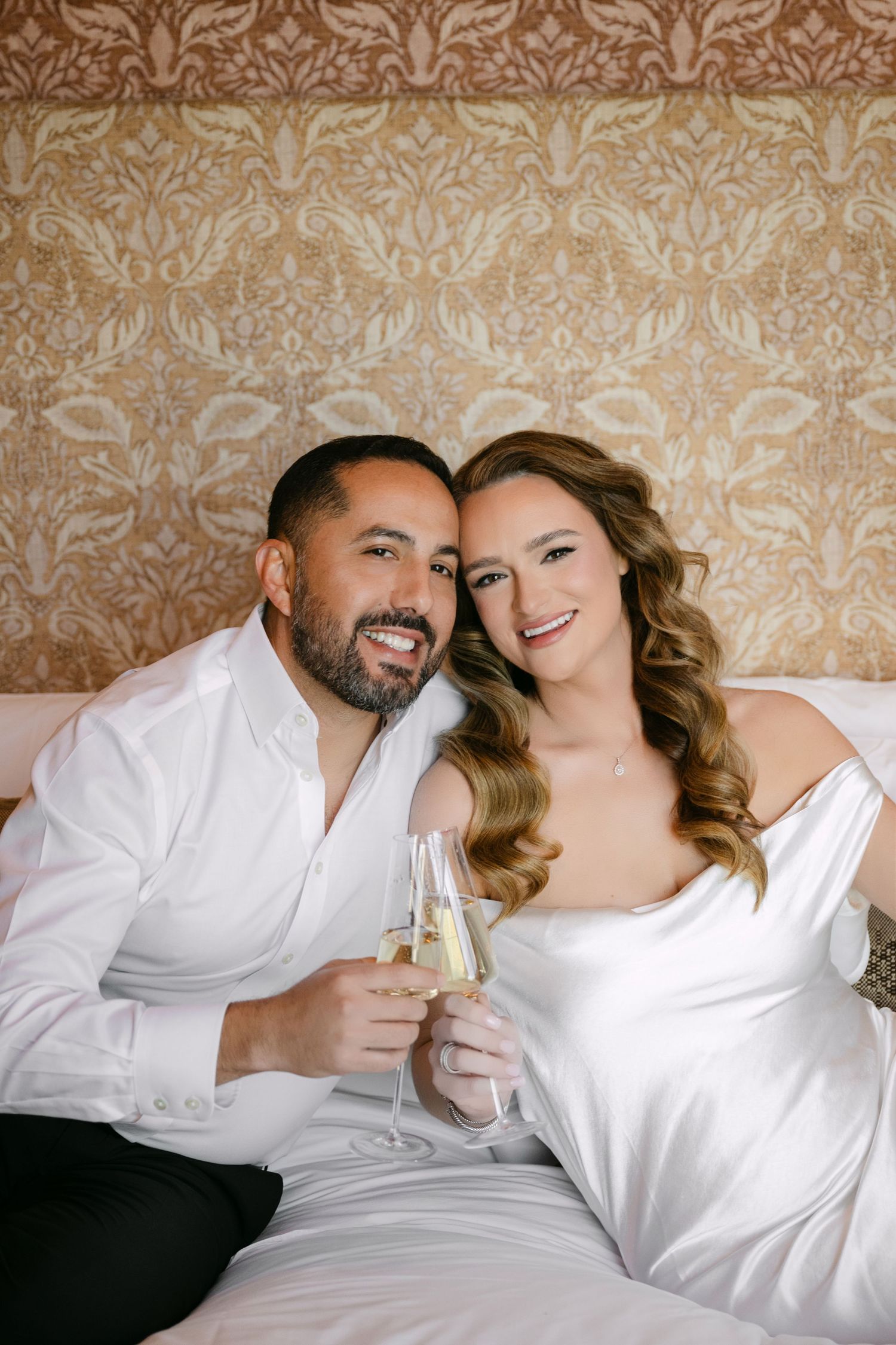 Happy newlyweds toasting with champagne glasses on a bed, smiling at camera in wedding attire.
