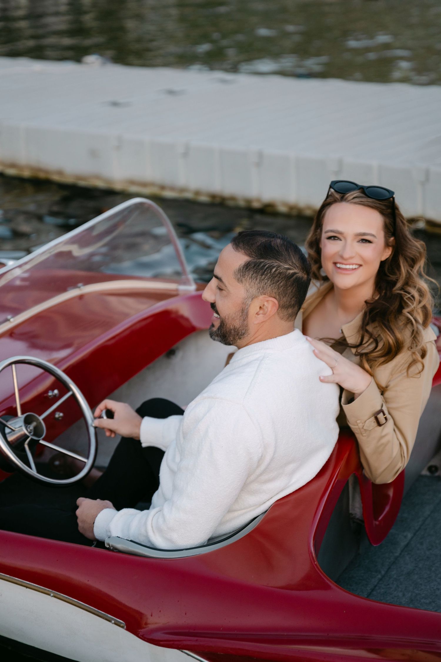 Smiling woman leans on mans shoulder as he steers a vintage red boat.