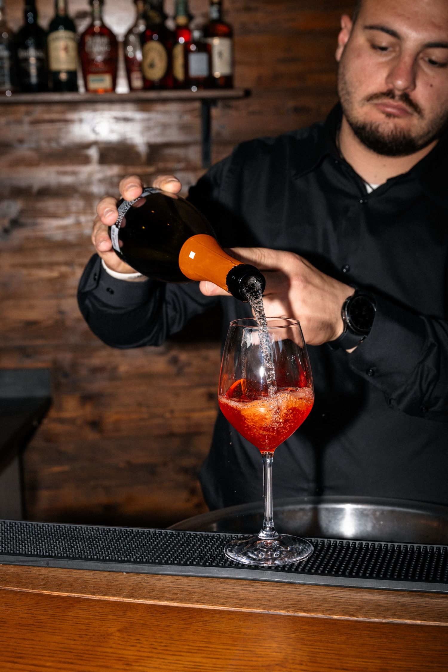 Bartender in black pouring Aperol Spritz cocktail into wine glass at rustic wooden bar.