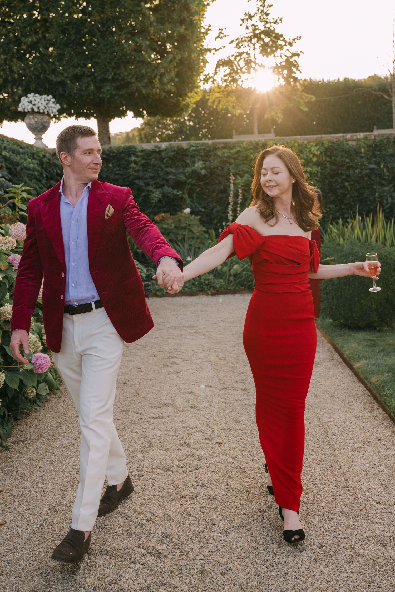 Couple walking hand in hand at sunset in garden, man in red velvet blazer, woman in red gown.