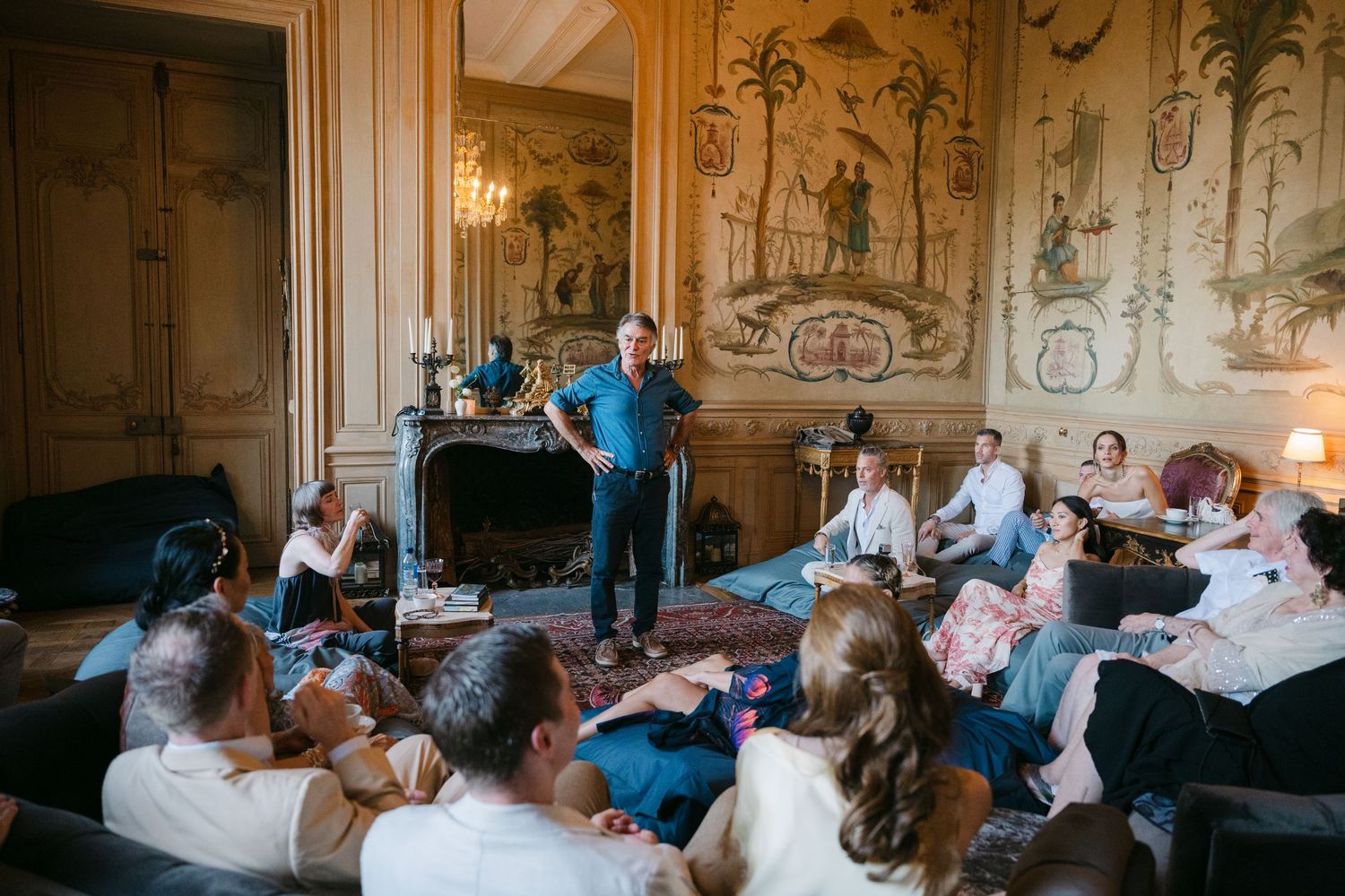 Man in blue shirt speaks to guests seated in an ornate French chateau salon with painted walls.