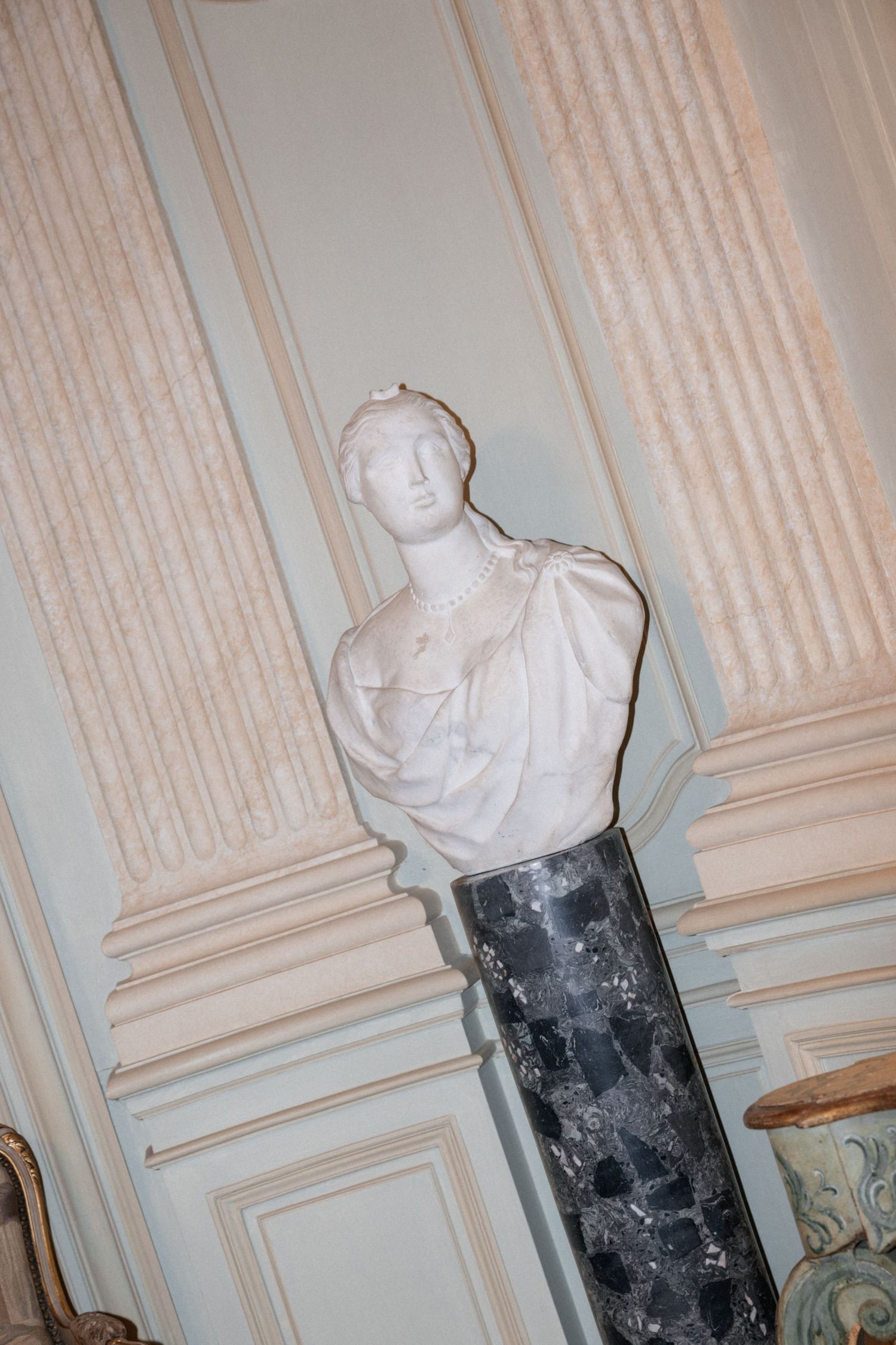 White marble classical bust sculpture on a dark marble pedestal in an ornate French chateau interior.