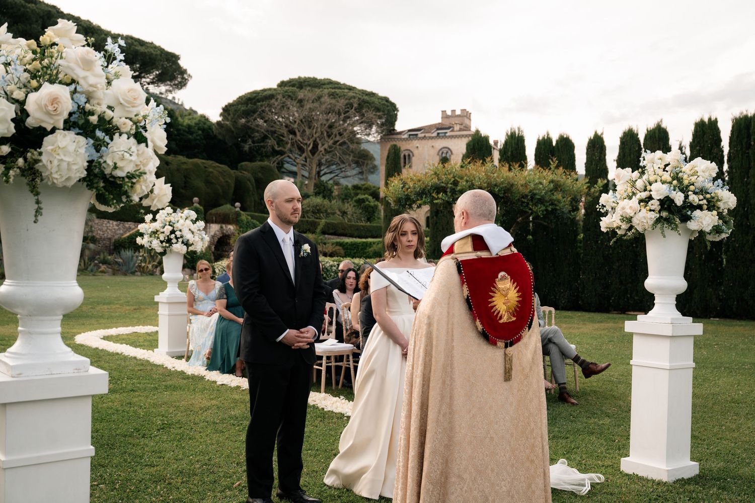 Couple exchanges vows at outdoor garden wedding ceremony with white floral arrangements.