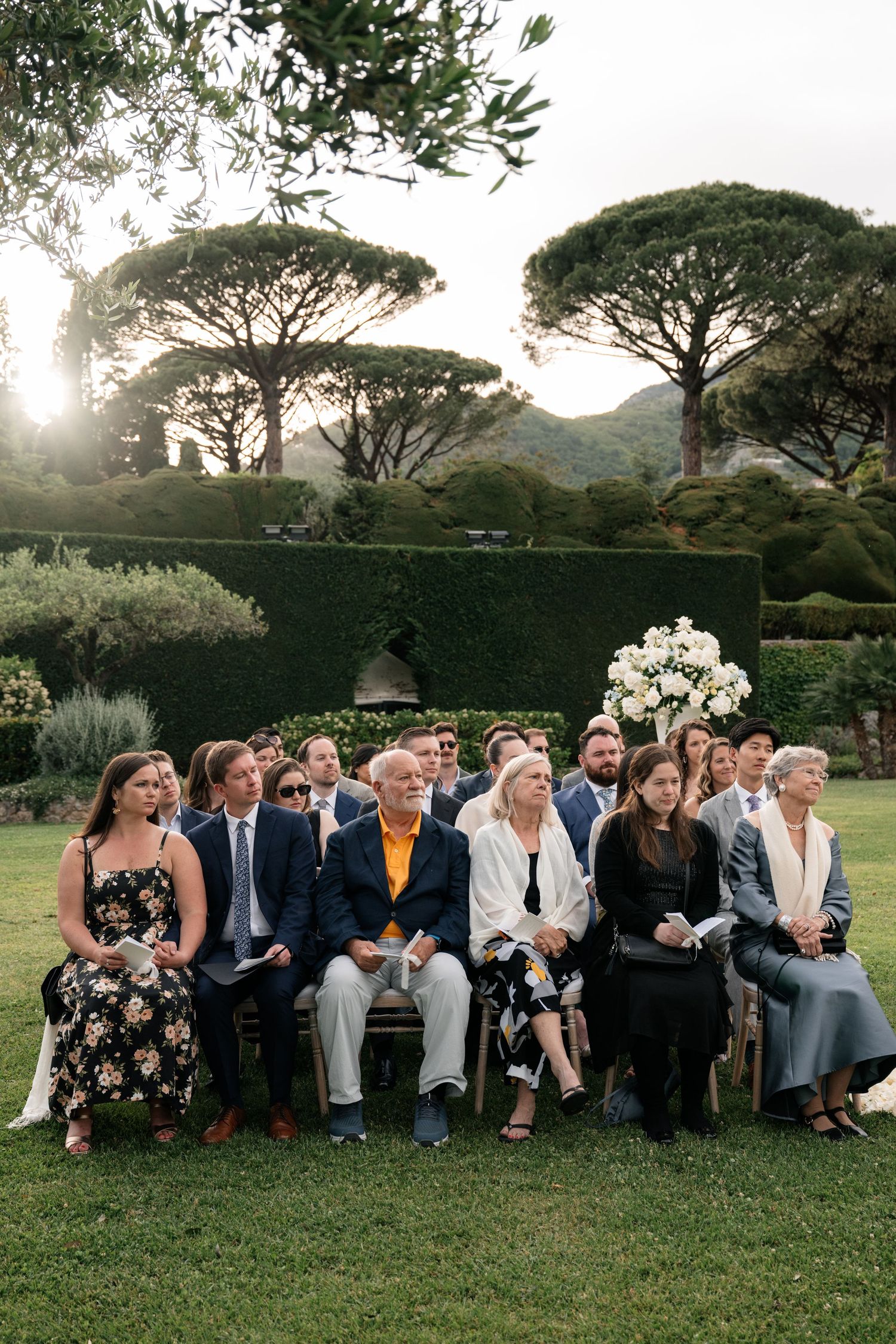 Wedding guests seated on lawn watching ceremony surrounded by manicured hedges and tall Italian pine trees.