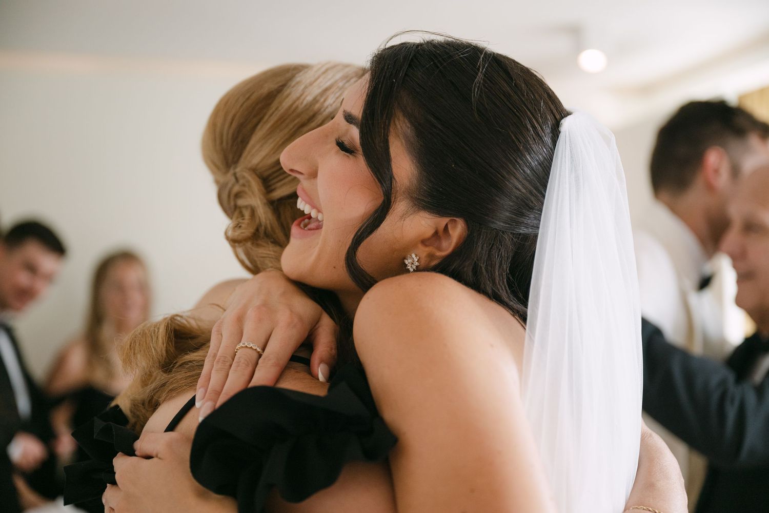 Bride in white veil shares warm embrace with blonde wedding guest after ceremony.