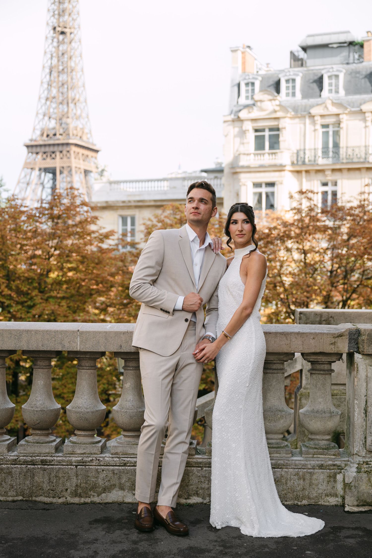 Elegant bride in white sequin gown and groom in beige suit pose on a stone balcony with the Eiffel Tower behind them.