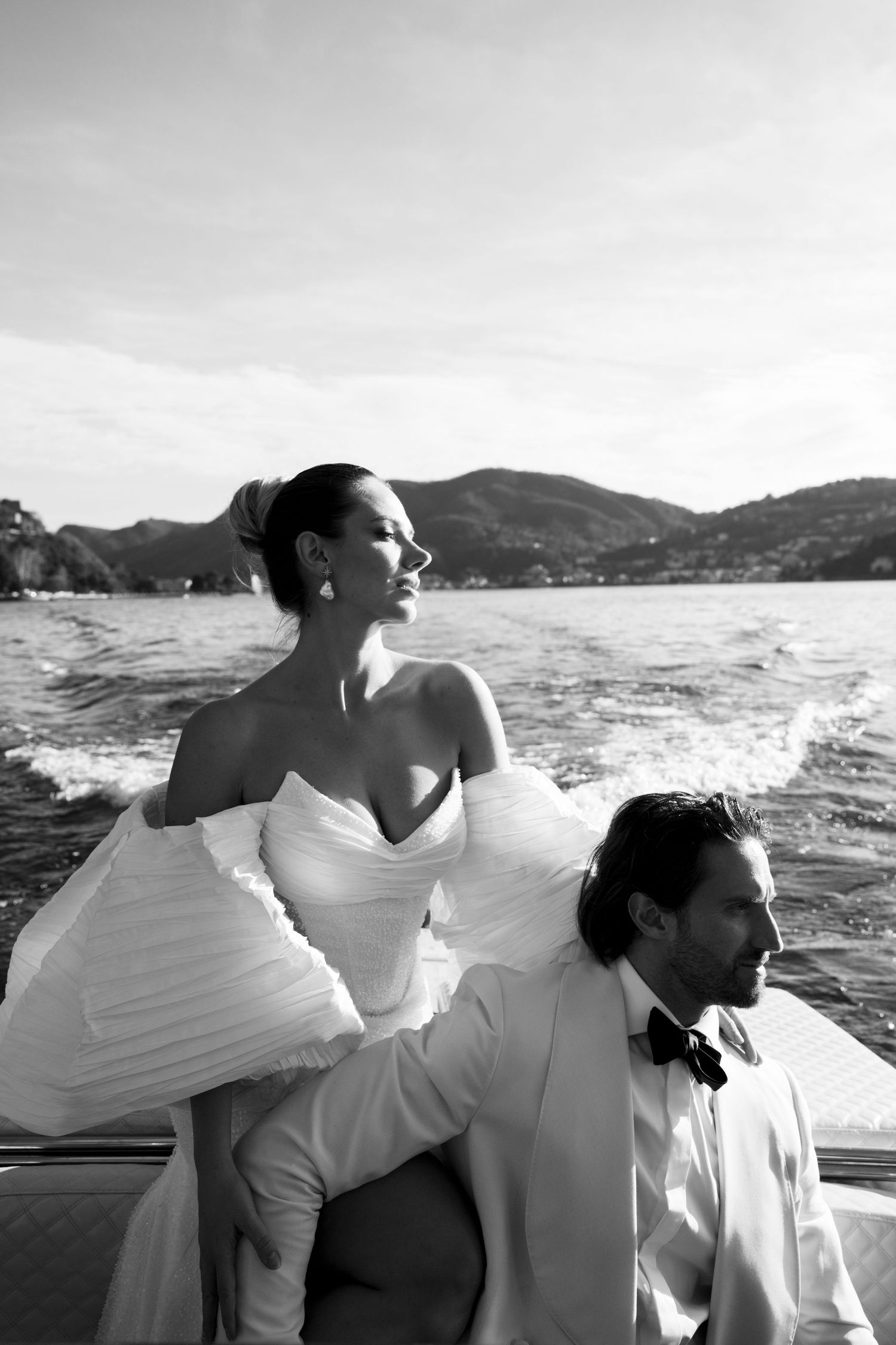 Black and white portrait of bride and groom on speedboat with Lake Como mountains in background.