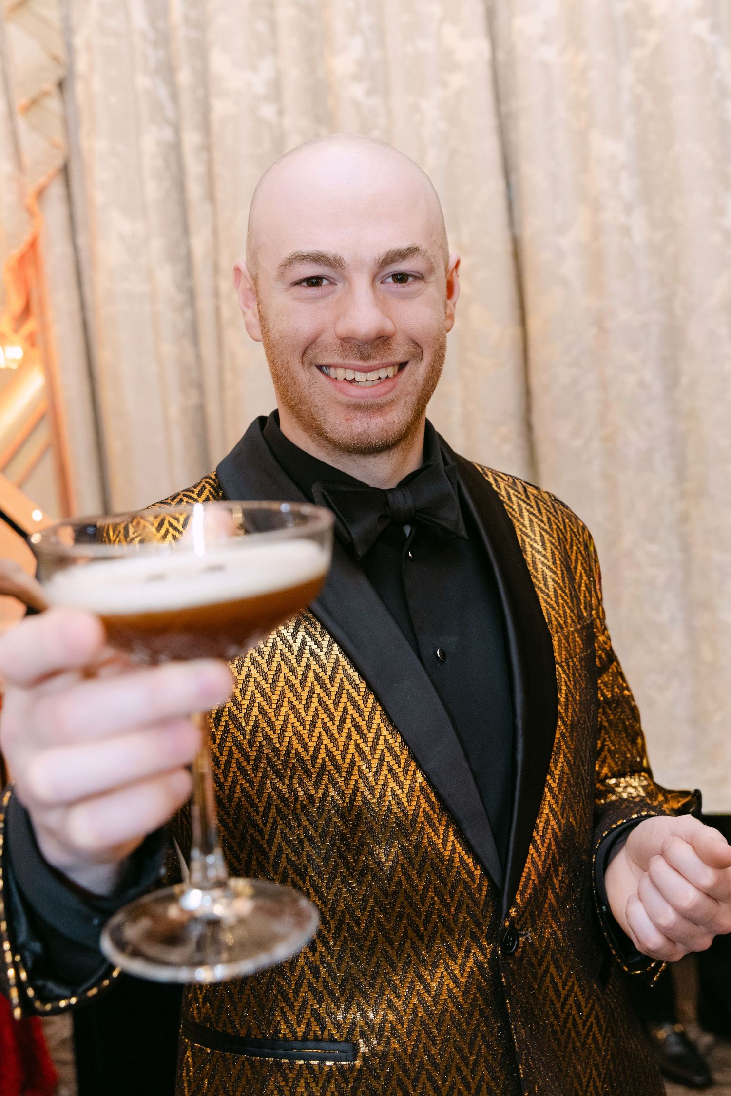 Smiling bald man in gold sequin tuxedo jacket toasting with espresso martini at elegant event.