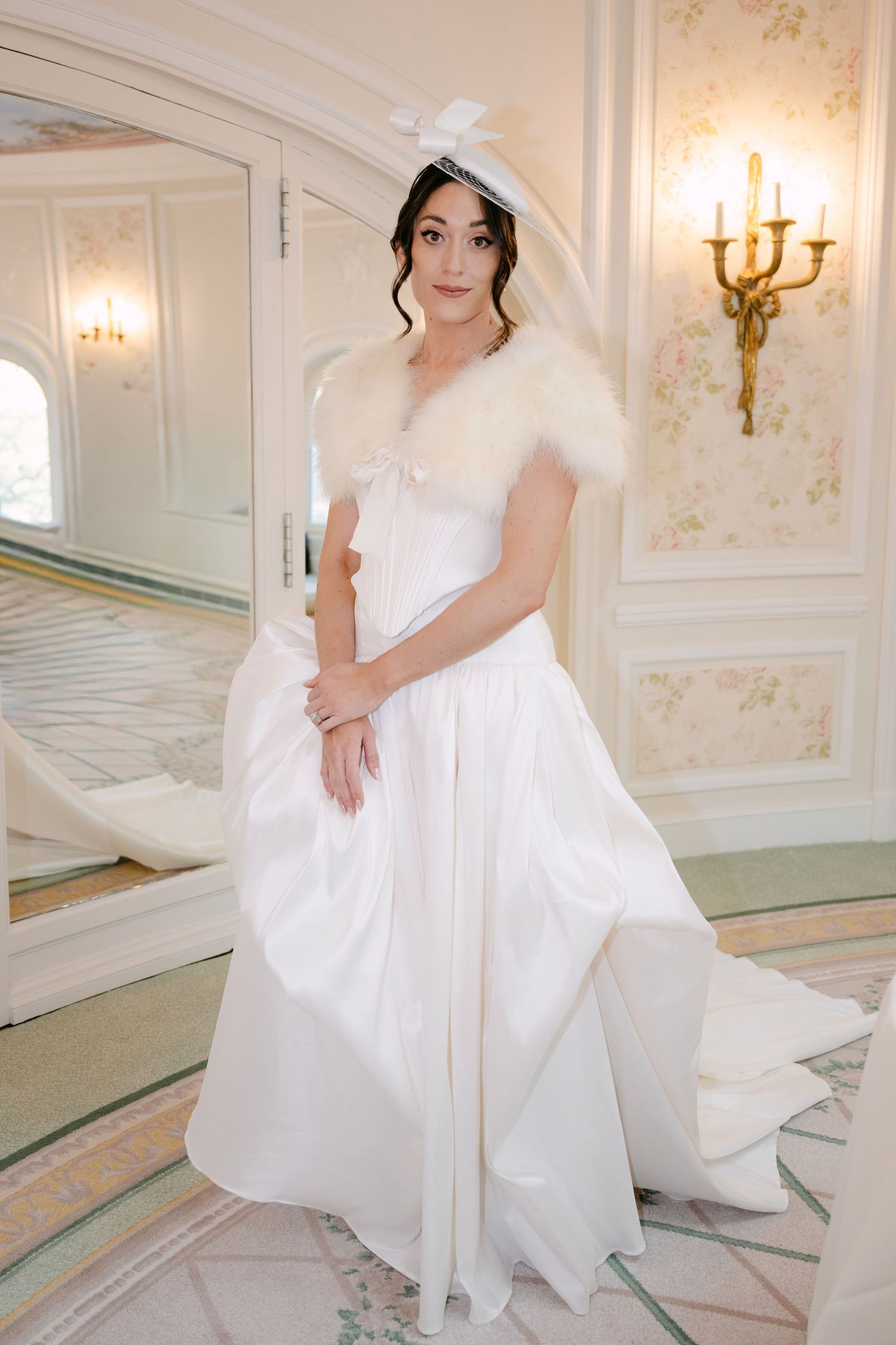 Bride in white satin ball gown with fur wrap and bow fascinator posing in ornate hotel suite.
