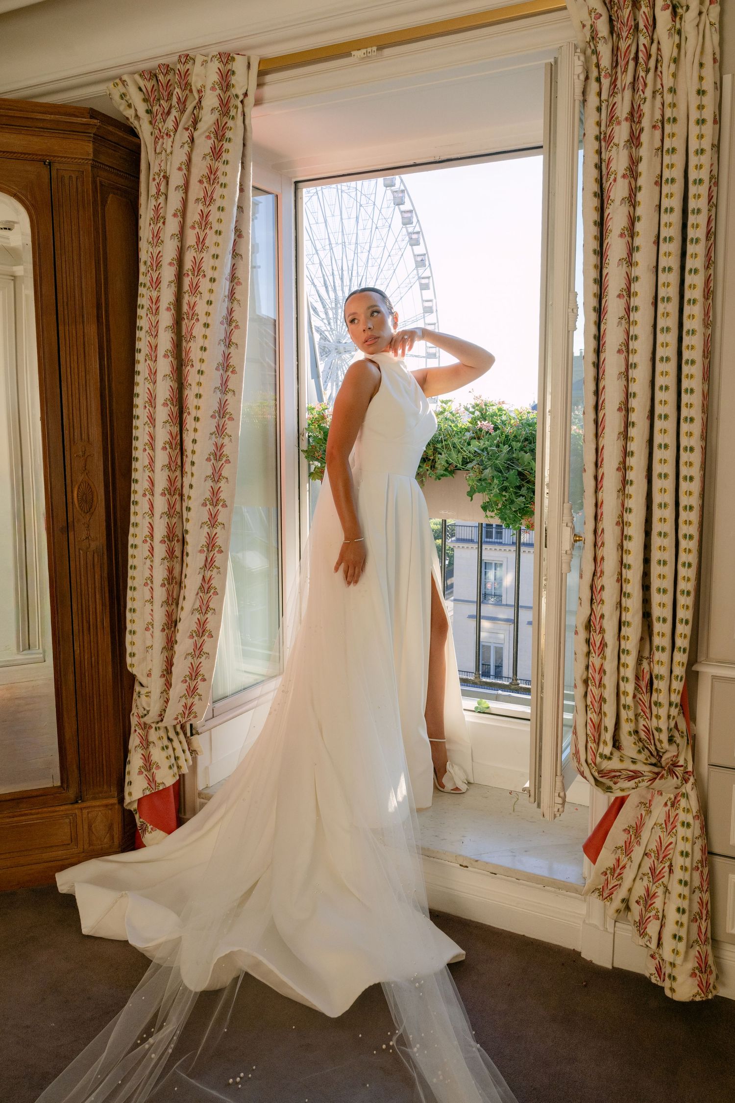 Bride in elegant white gown with train stands in Parisian window with Ferris wheel view.