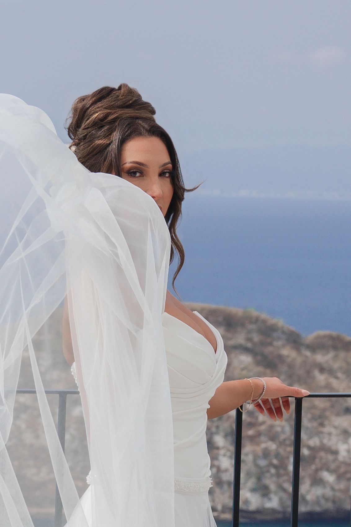 Bride in white gown and flowing veil poses on balcony with ocean and rocky cliffs in background.