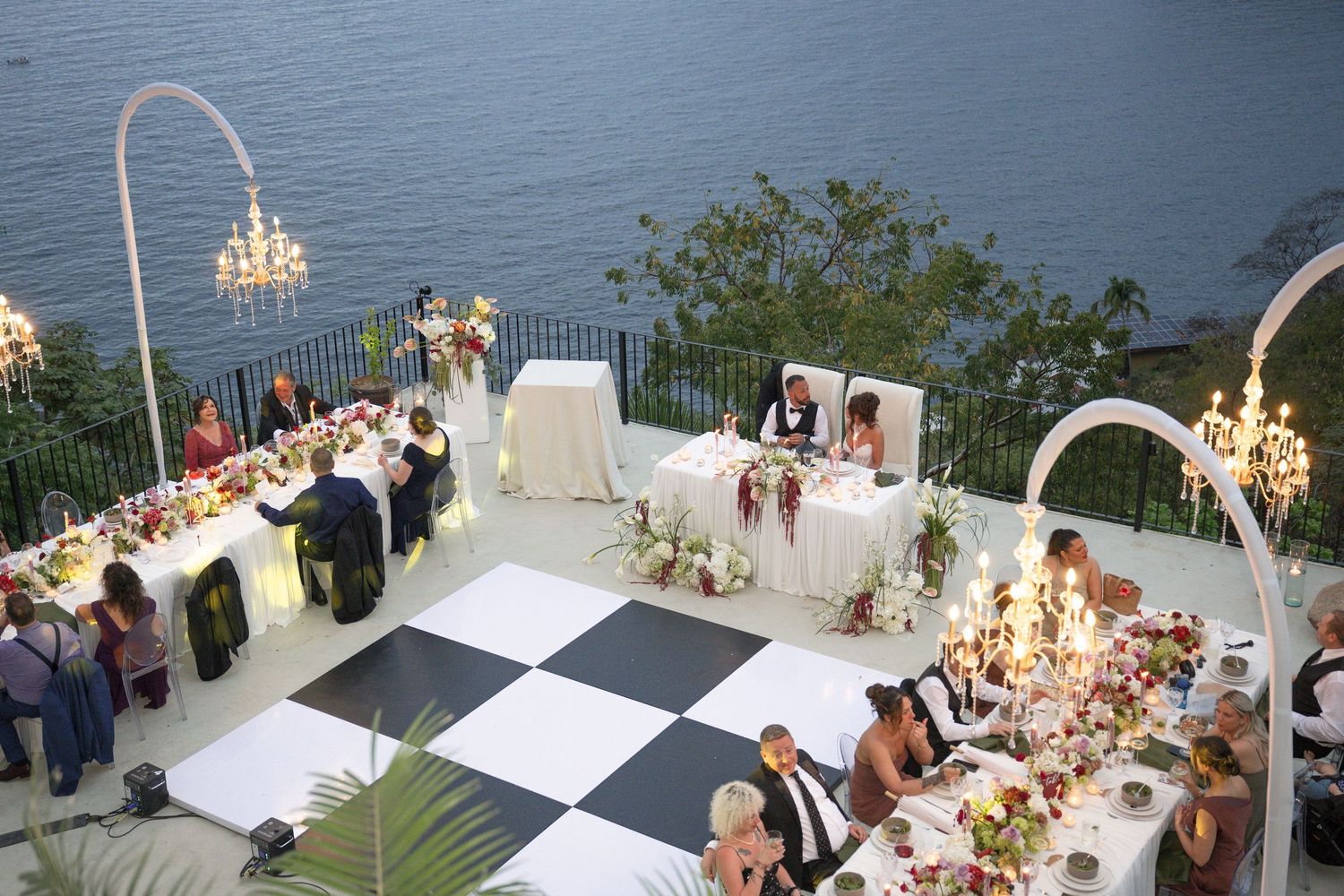 Elegant outdoor wedding reception with checkered dance floor, long decorated tables, and chandeliers overlooking a scenic waterfront.