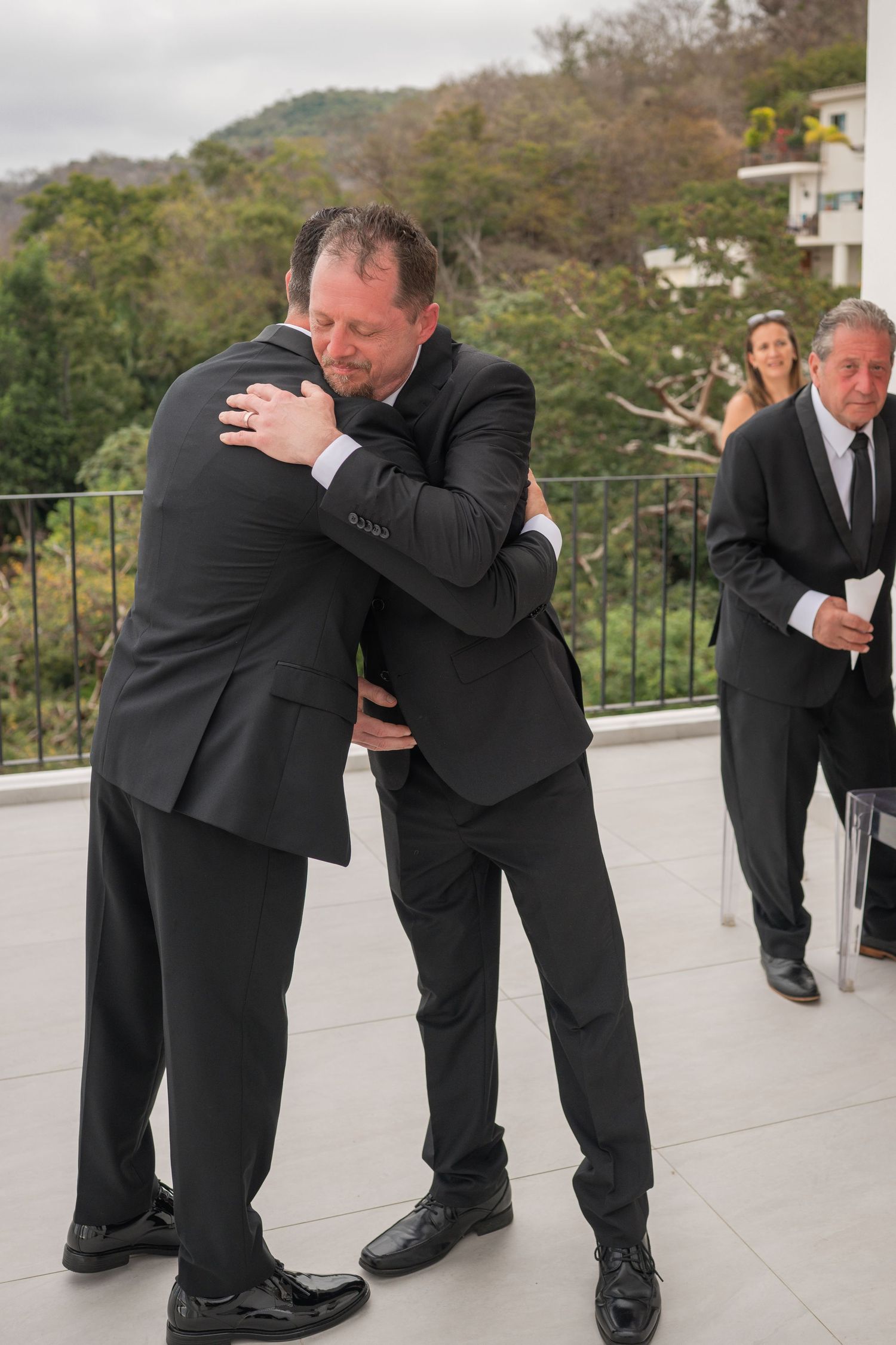 Two men in black suits share an emotional hug during a wedding ceremony on an outdoor terrace.